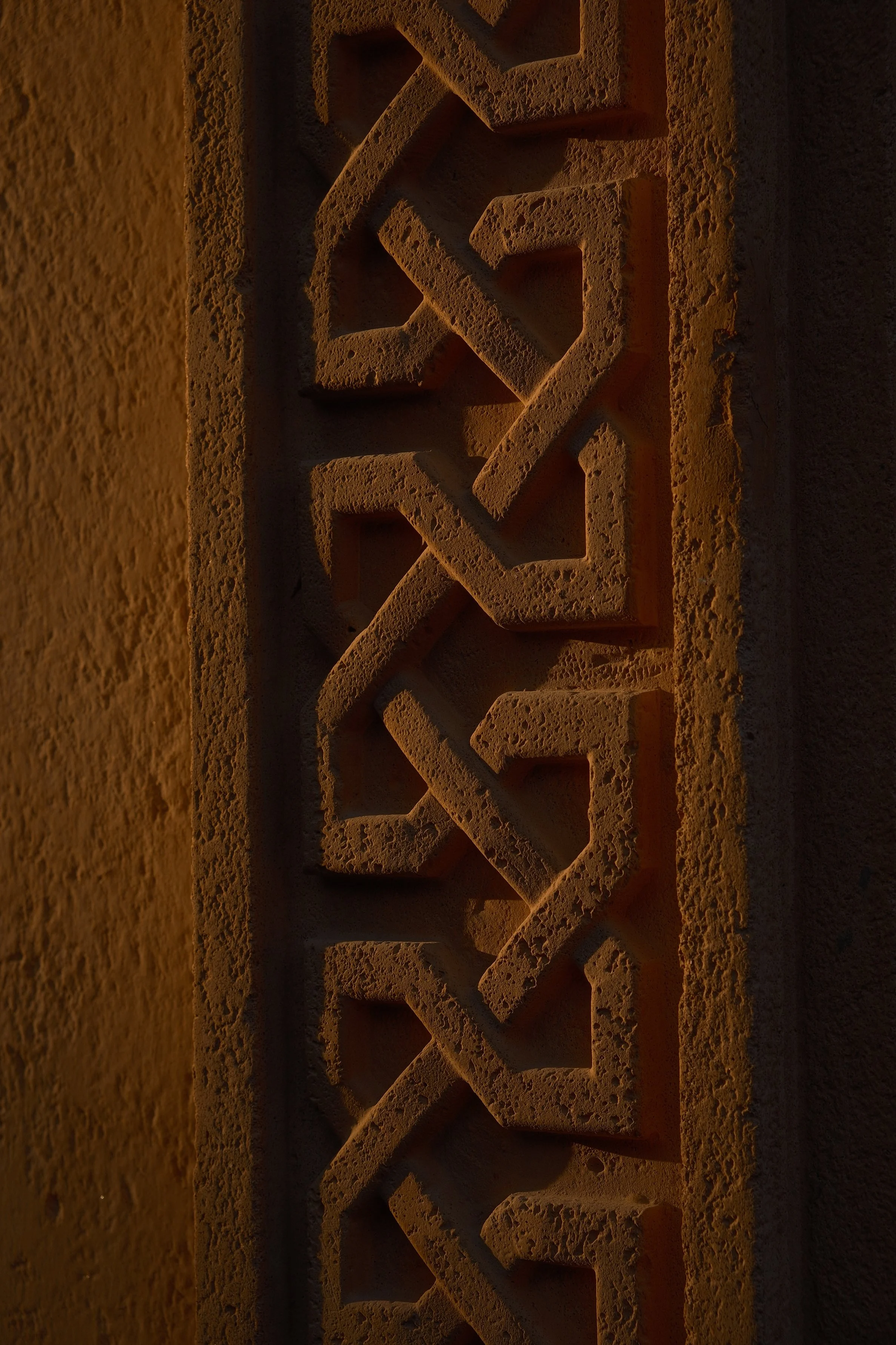 Close-up of a decorative terracotta wall panel with intricate geometric patterns and textured surface, illuminated by warm light.