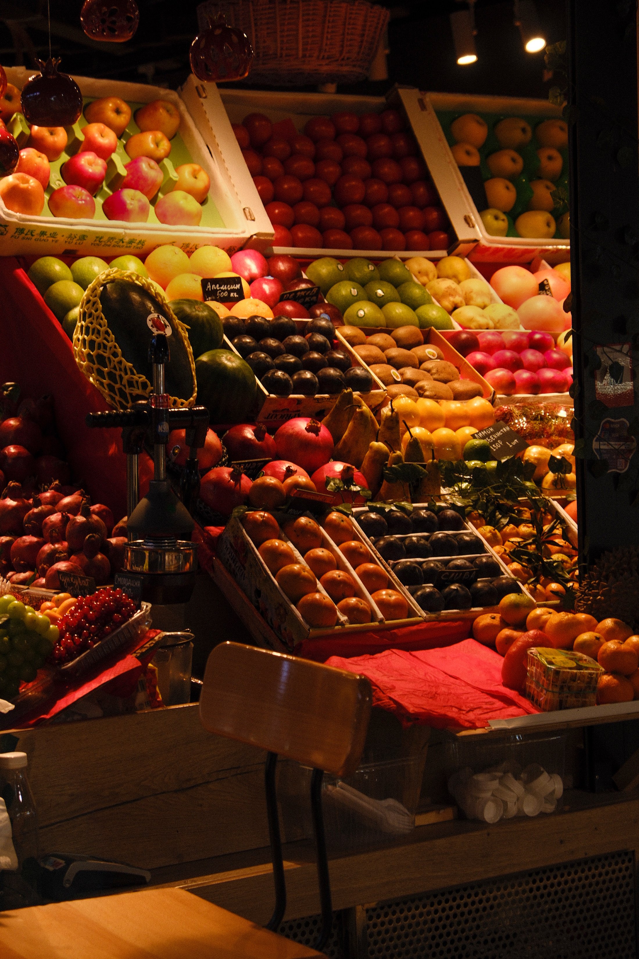 Display of various fresh fruits on a market stall, including apples, tomatoes, watermelons, peaches, plums, and grapes, arranged in baskets and boxes under warm lighting.