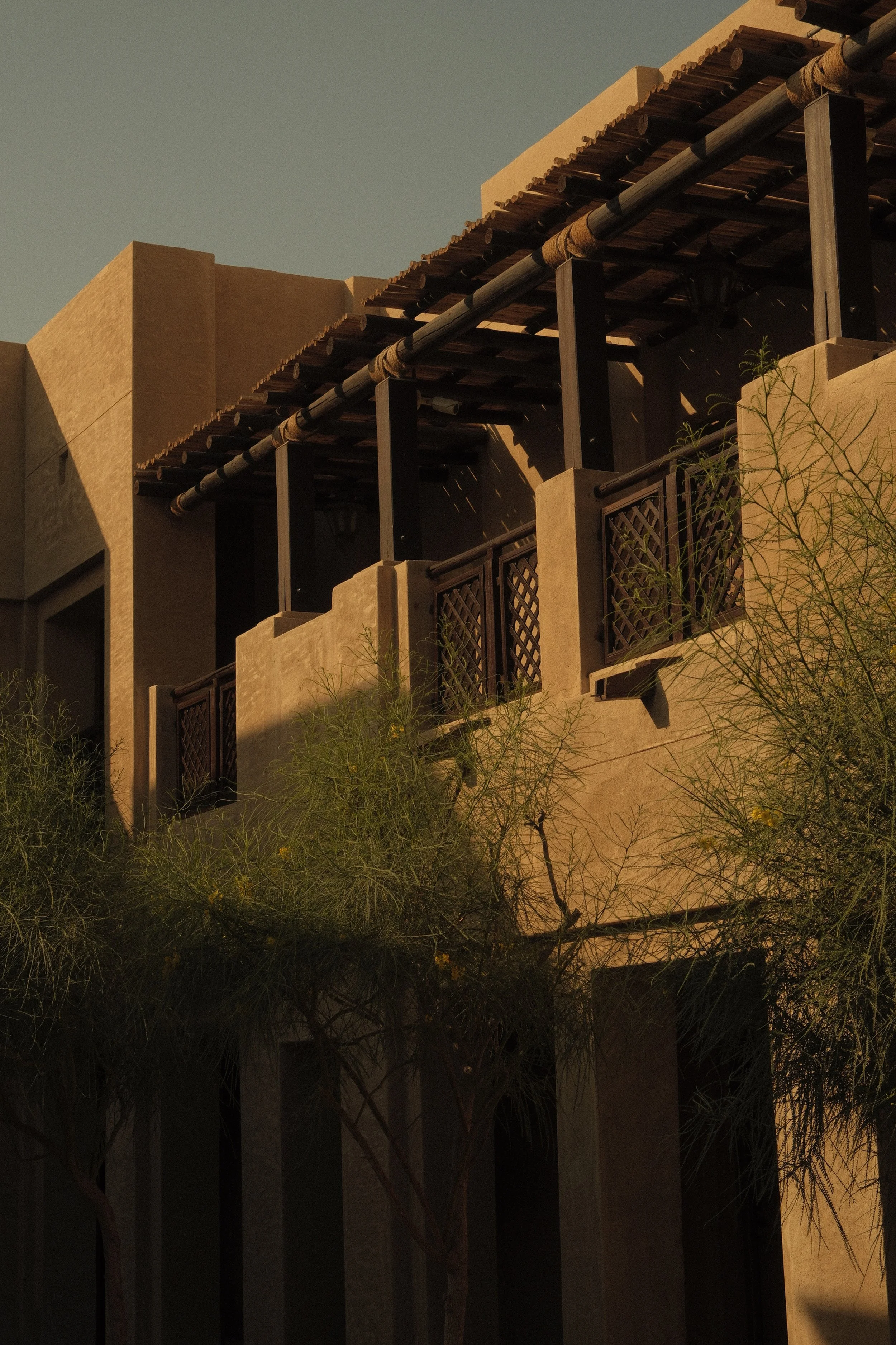 A balcony with a wooden roof, black railing, and stucco walls, with desert plants in front and the sky in the background.