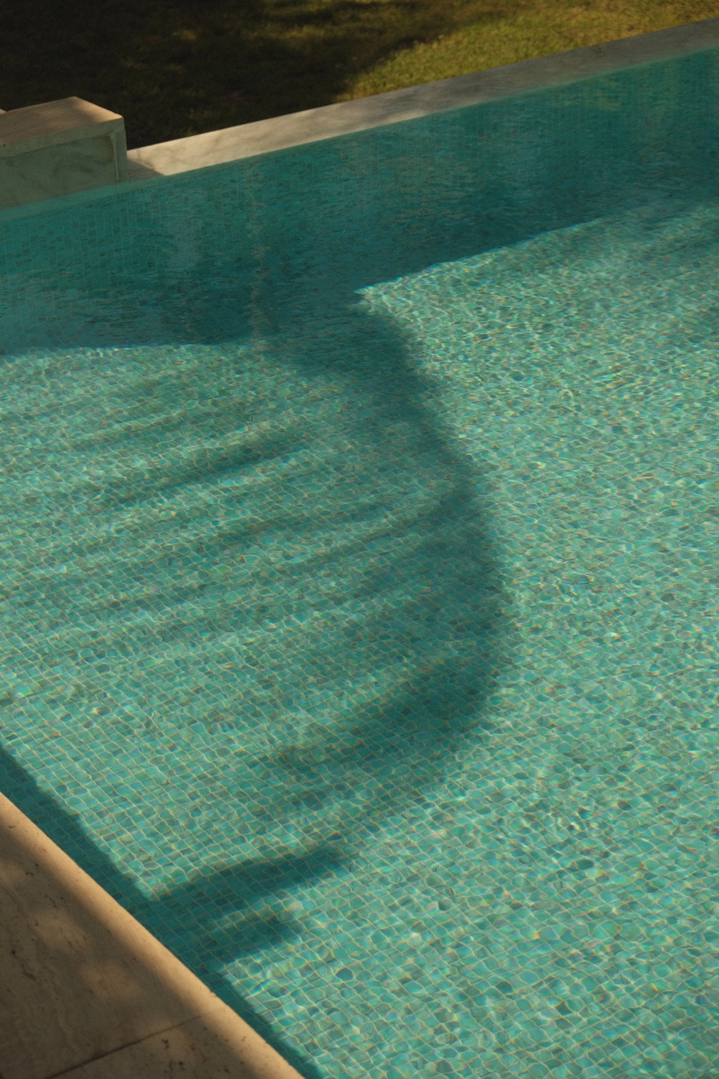 Close-up of a swimming pool with clear water and small blue tiles, surrounded by a stone or concrete edge, with a grassy area in the background.