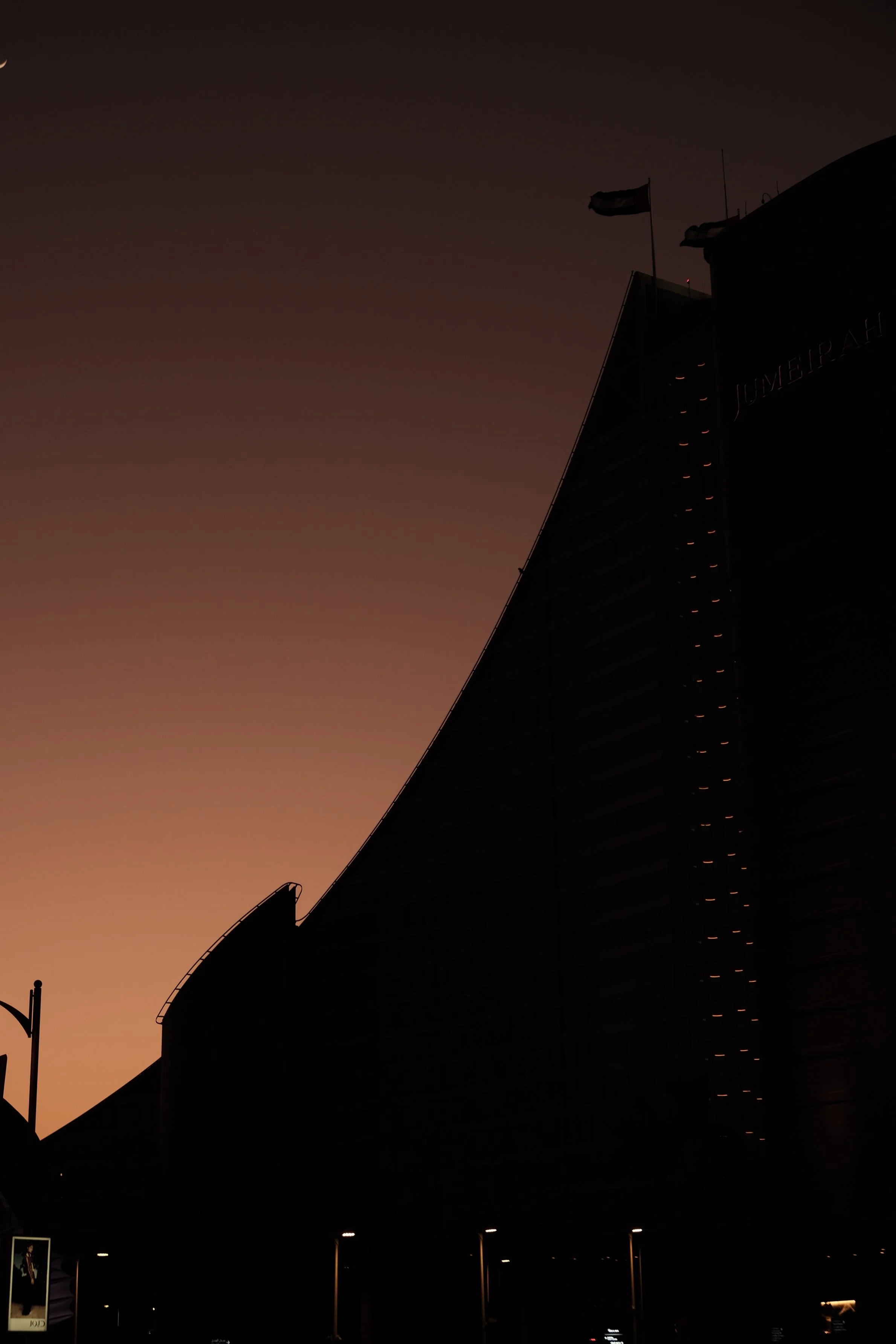 Dark silhouette of a modern building with flags on top against a dusky orange and purple sky.