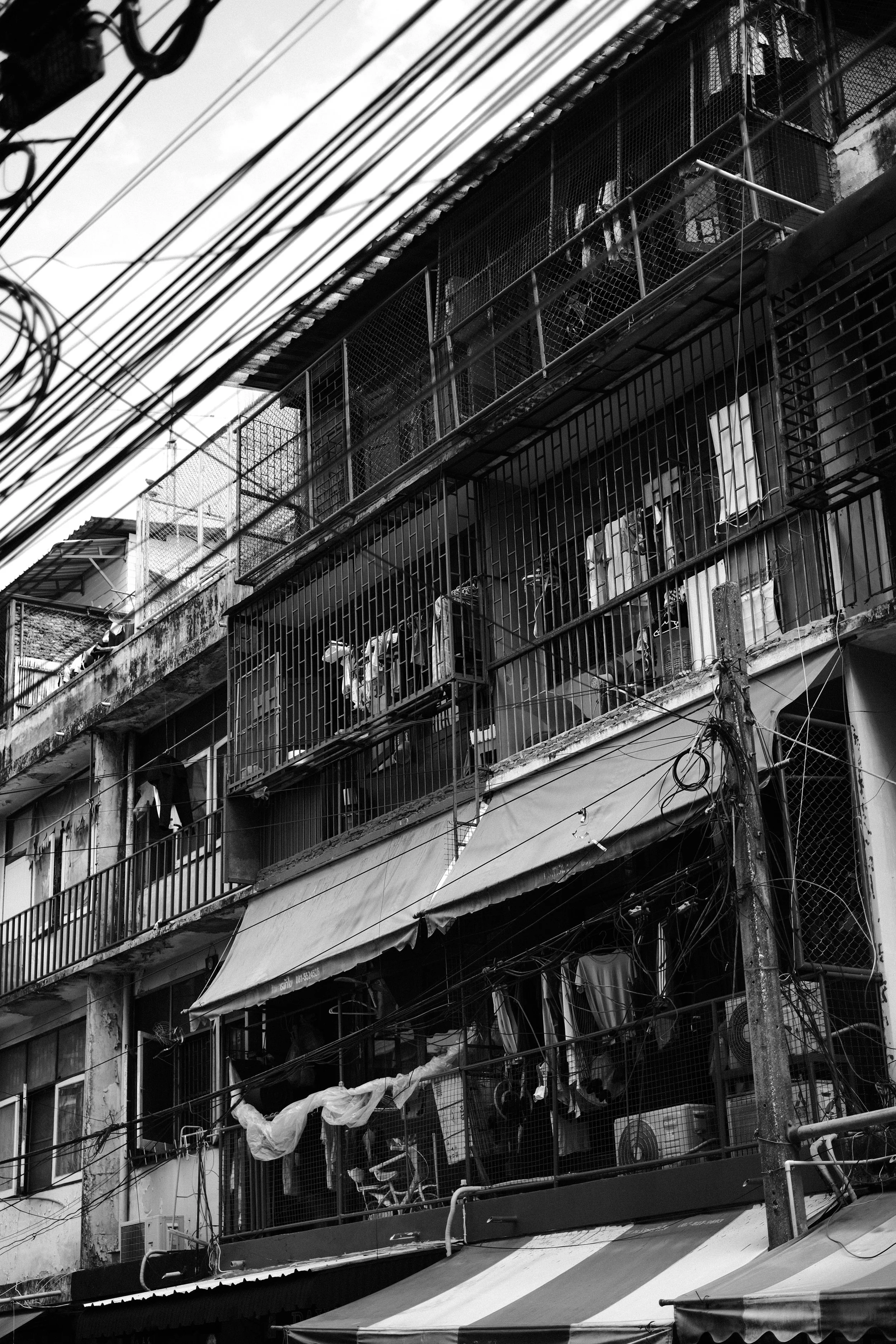 Black and white photo of a multi-story building with balconies, laundry hanging, and multiple electrical wires in the foreground.