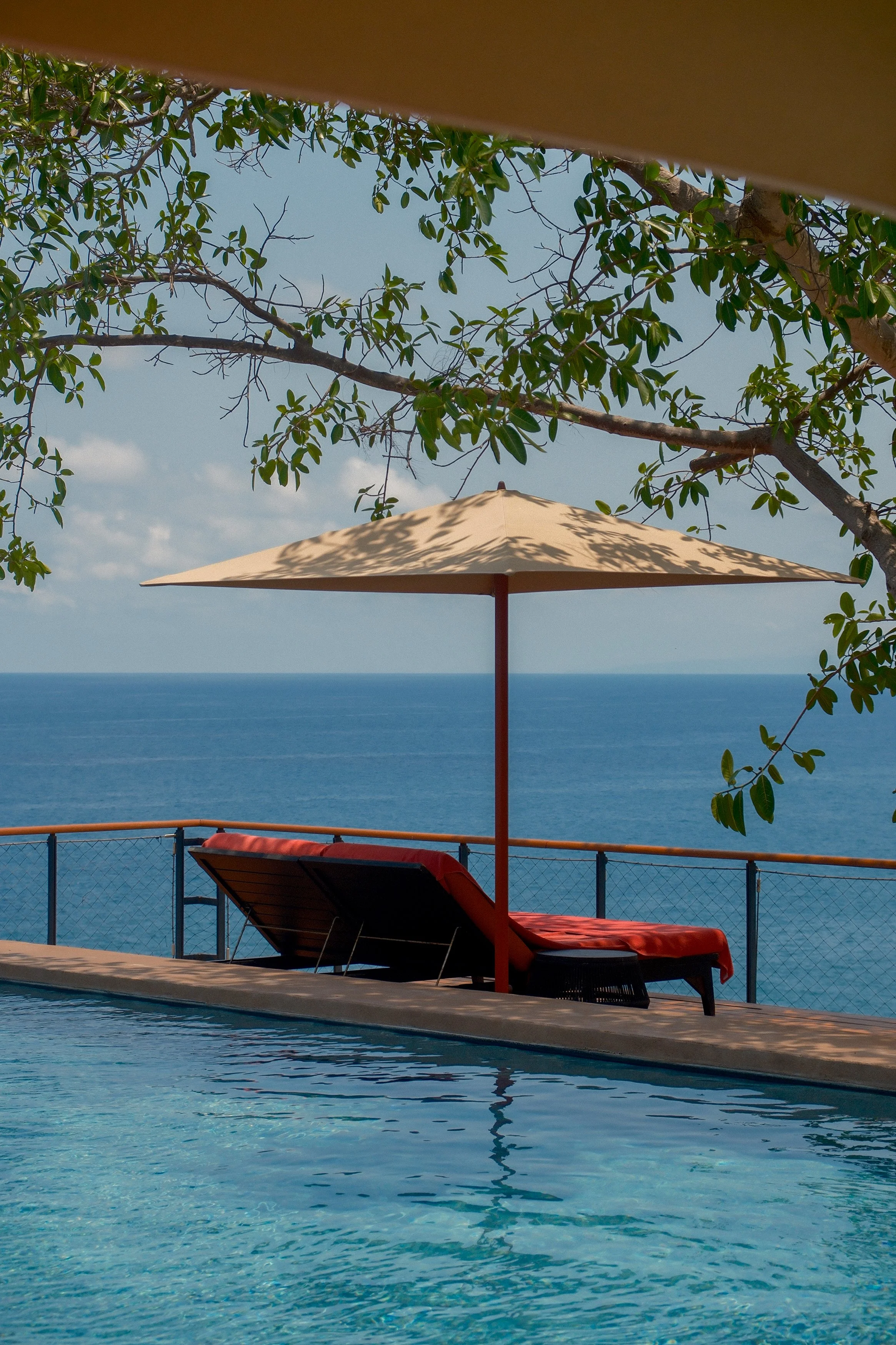 A view of a swimming pool with a single lounge chair covered with a red towel, shaded by a beige umbrella. In the background, there is a fence, a large tree with green leaves, and the ocean under a blue sky.