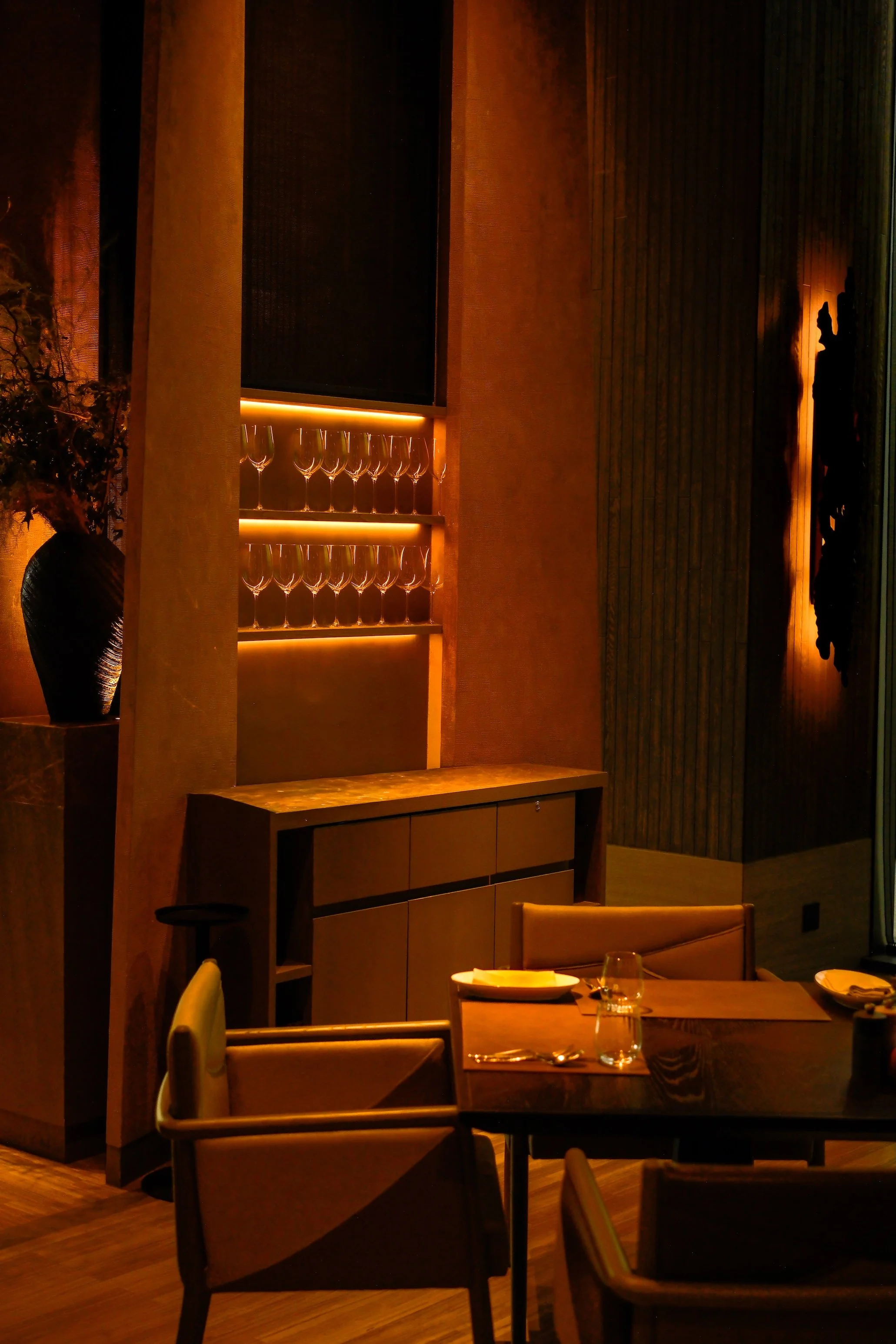 A dimly lit restaurant interior with wooden walls, a display of wine glasses on lit shelves, a vase with flowers, and tables set with plates, glasses, and silverware.
