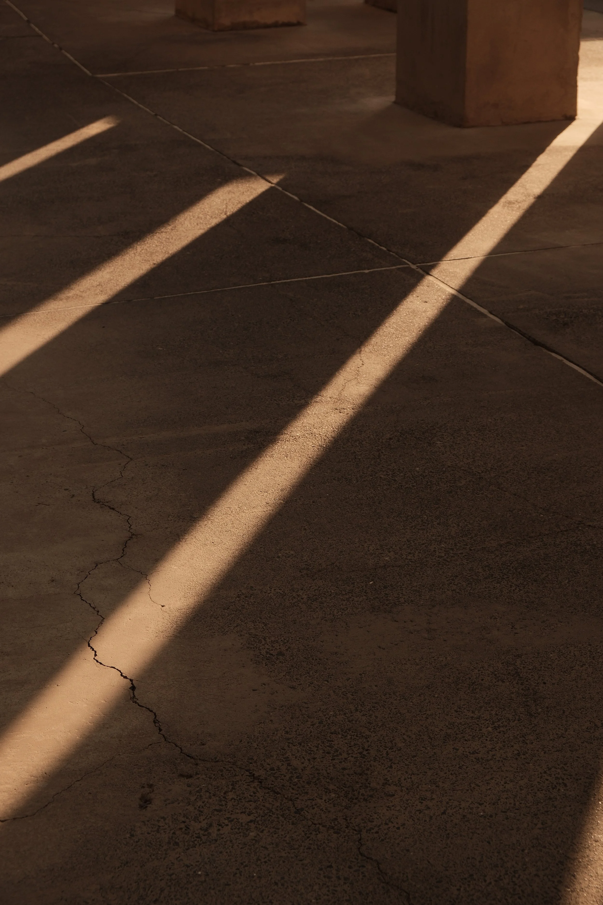 Sunlight casts long shadows across a cracked concrete sidewalk near supporting columns made of brick or stone.
