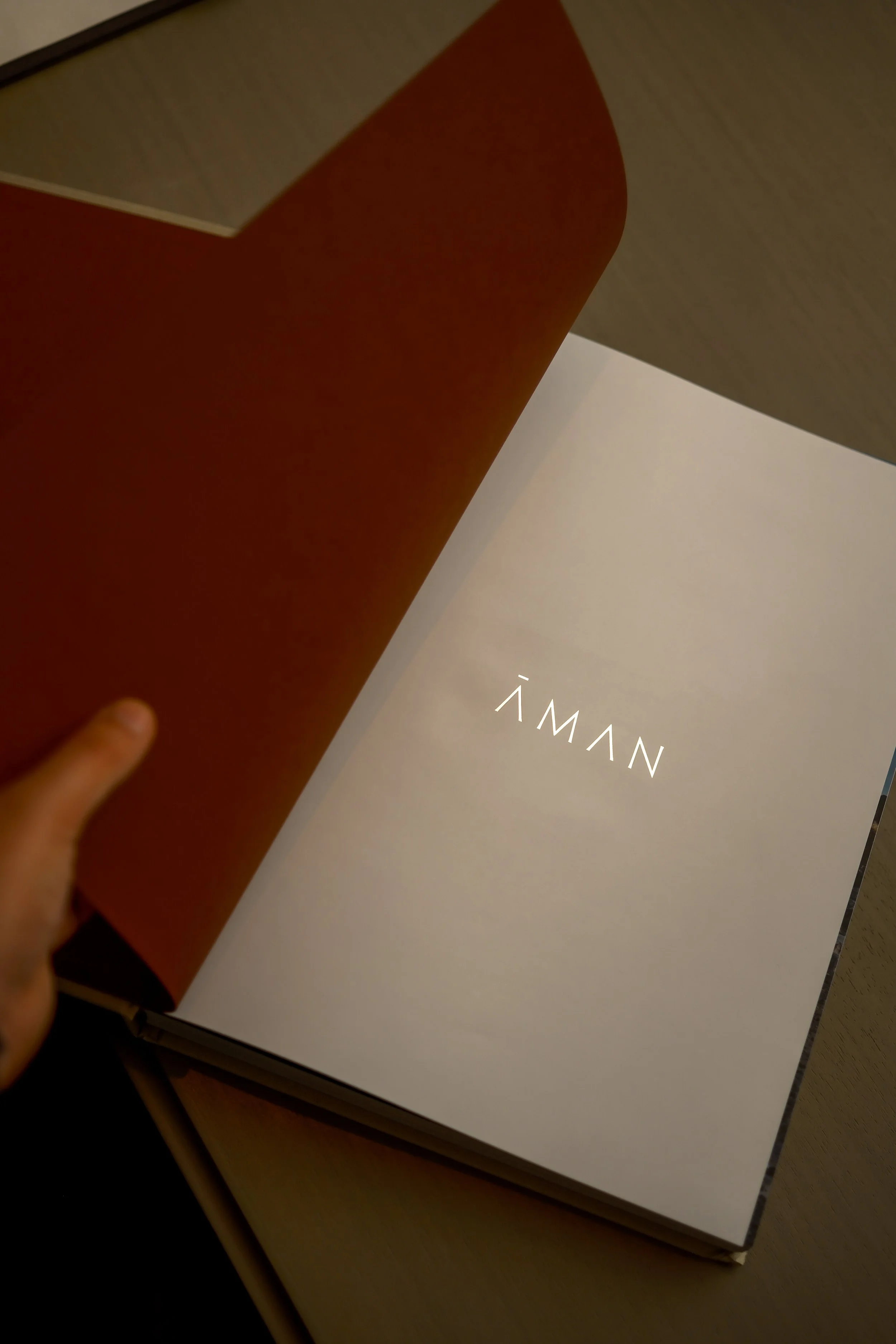 Close-up of a person holding a red hardcover book or box, with a white page or cover underneath that has the word 'NĀWALY' written on it.