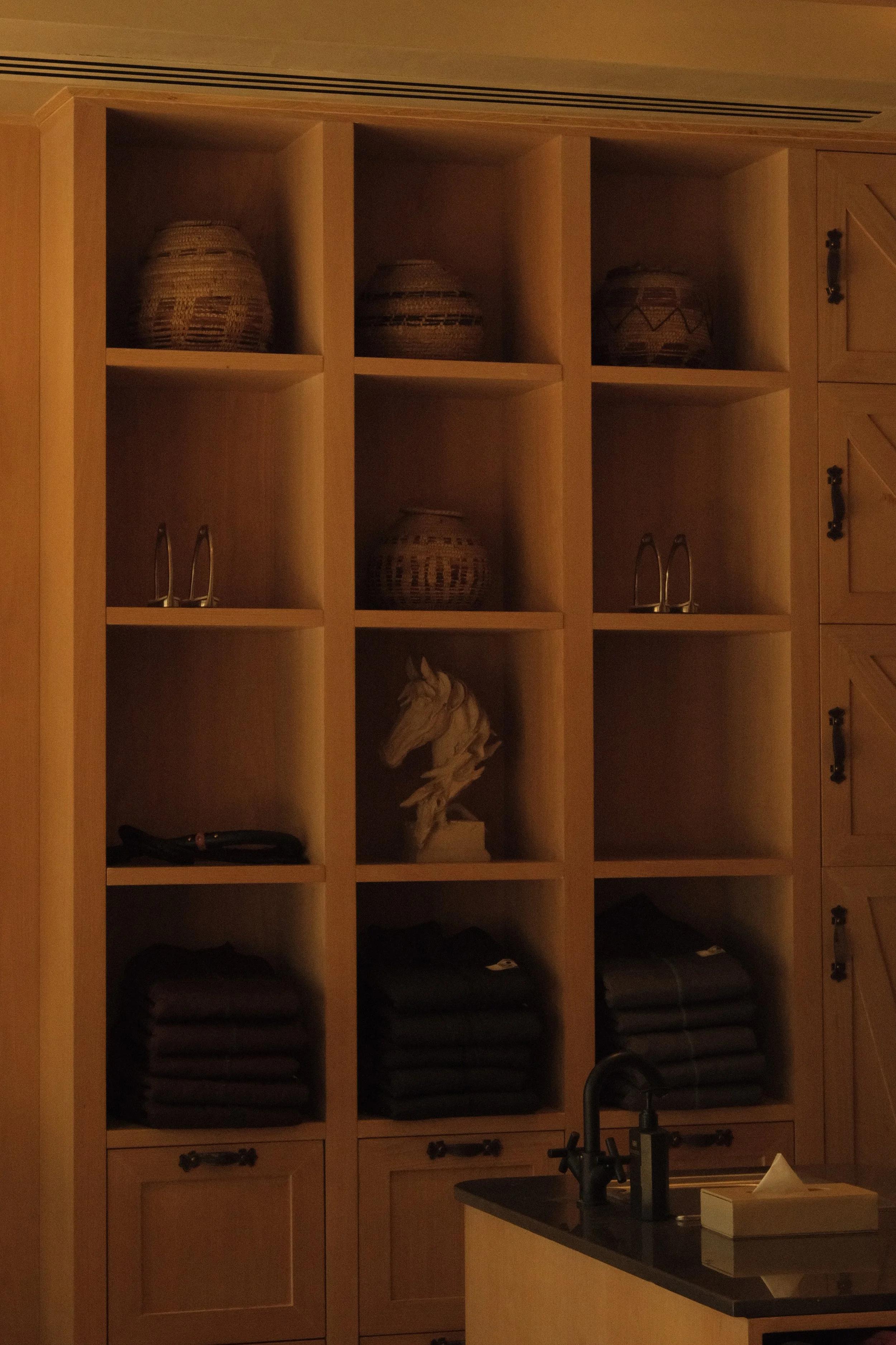 Wooden kitchen cabinet with open cubby shelves containing woven baskets, a decorative horse figurine, and folded dark-colored towels. A kitchen sink with a black faucet is in the foreground.