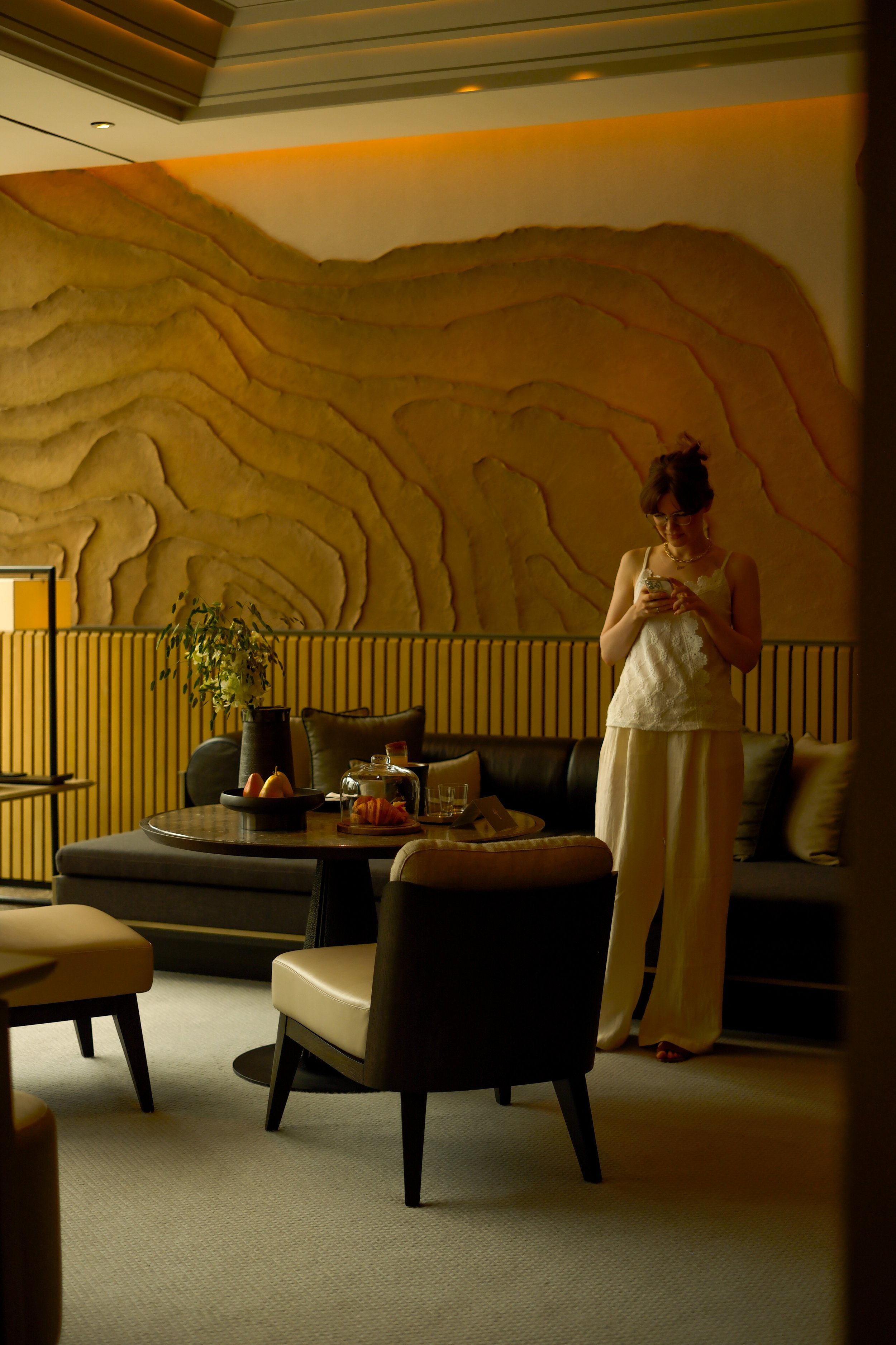 A woman standing in a modern, stylish restaurant or lounge, looking at her phone. The interior features a large abstract wall art, a round table with a black fruit bowl and a glass dome, and various chairs and sofas. The setting is cozy with warm lig