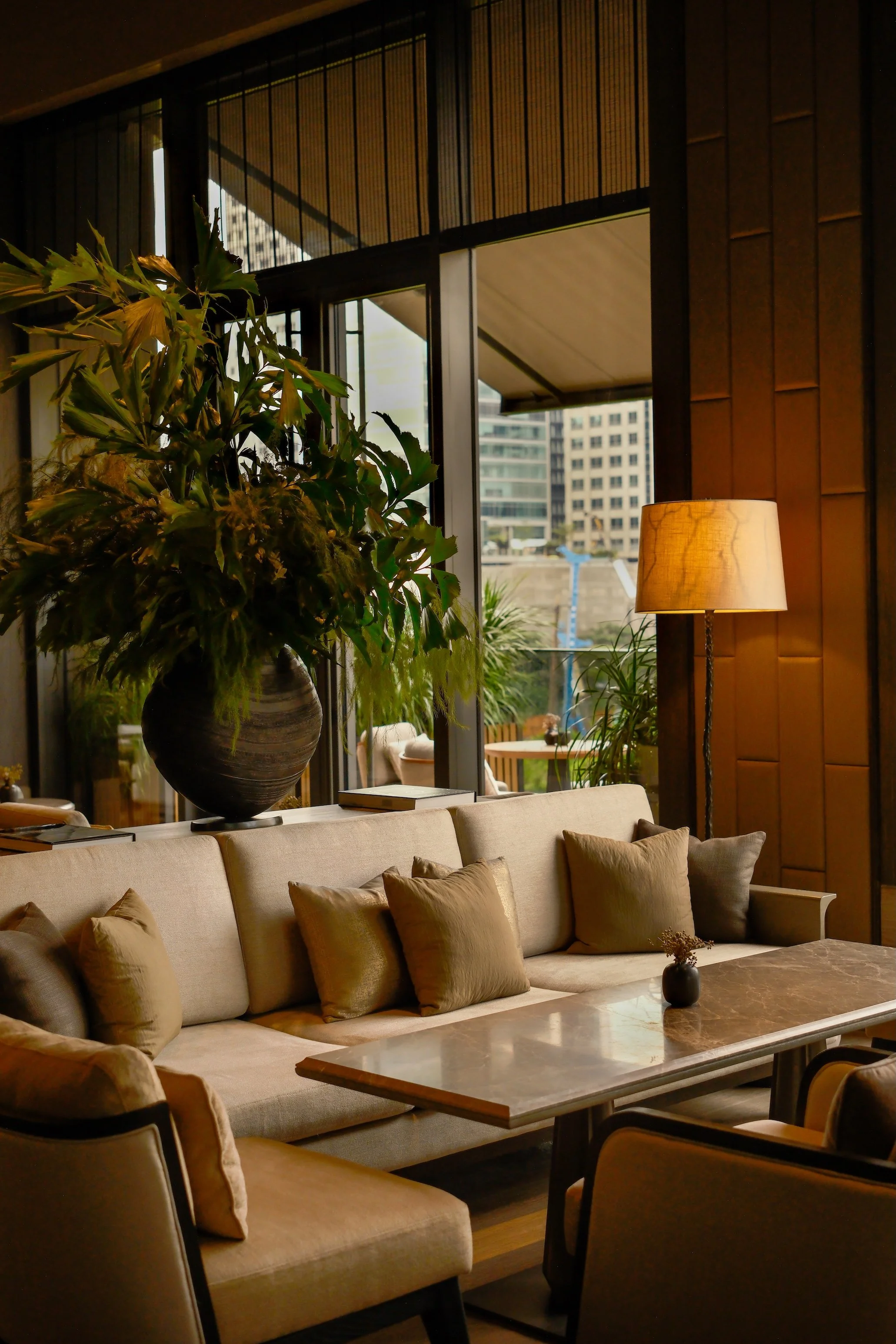 Interior of a modern lounge or hotel lobby with beige sofa and cushions, large potted plant, marble coffee table, table lamp, and large windows revealing city buildings outside.