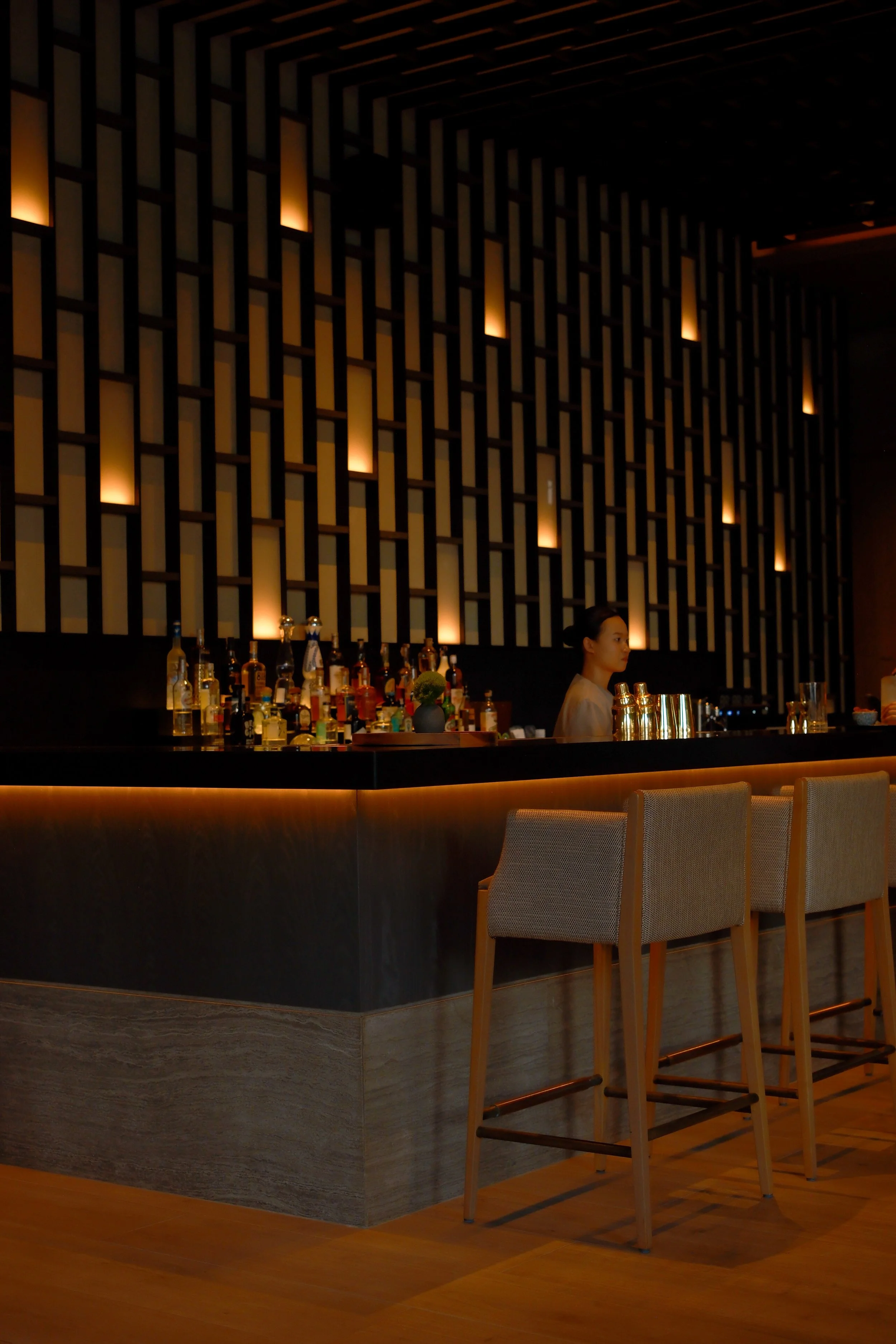 A modern hotel bar with a textured wall featuring geometric lighting, a bar counter with various bottles, and bar stools with beige upholstery and light wooden legs.