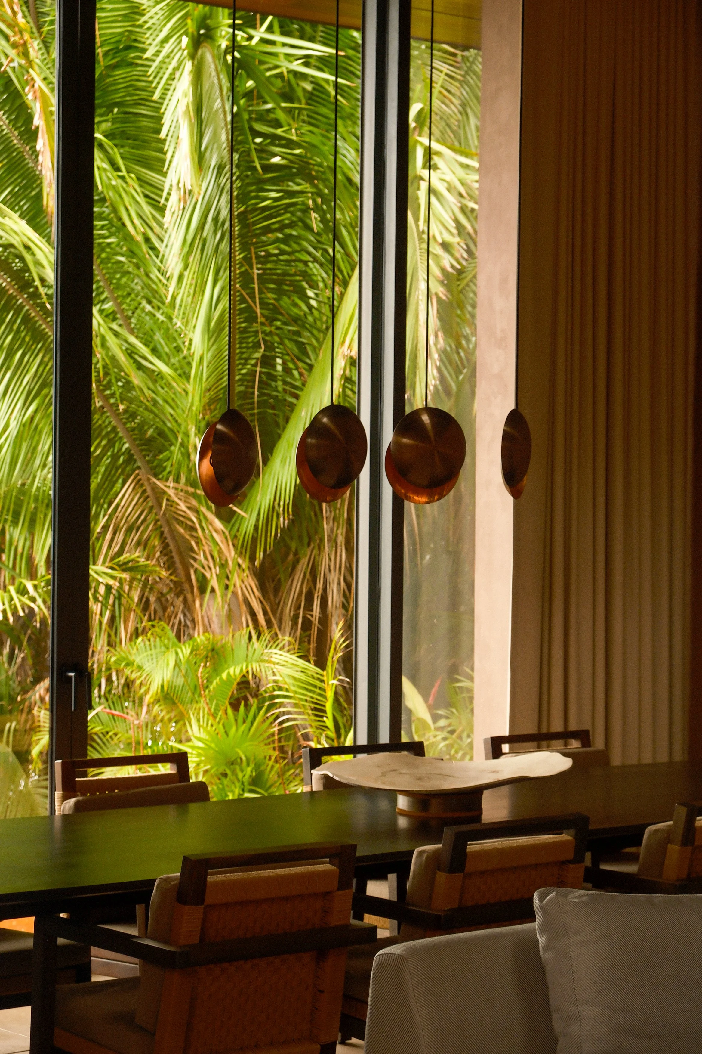 Interior of a modern dining area with wooden chairs around a dark table, large floor-to-ceiling windows showing lush green tropical plants outside, and decorative hanging copper bowls inside.