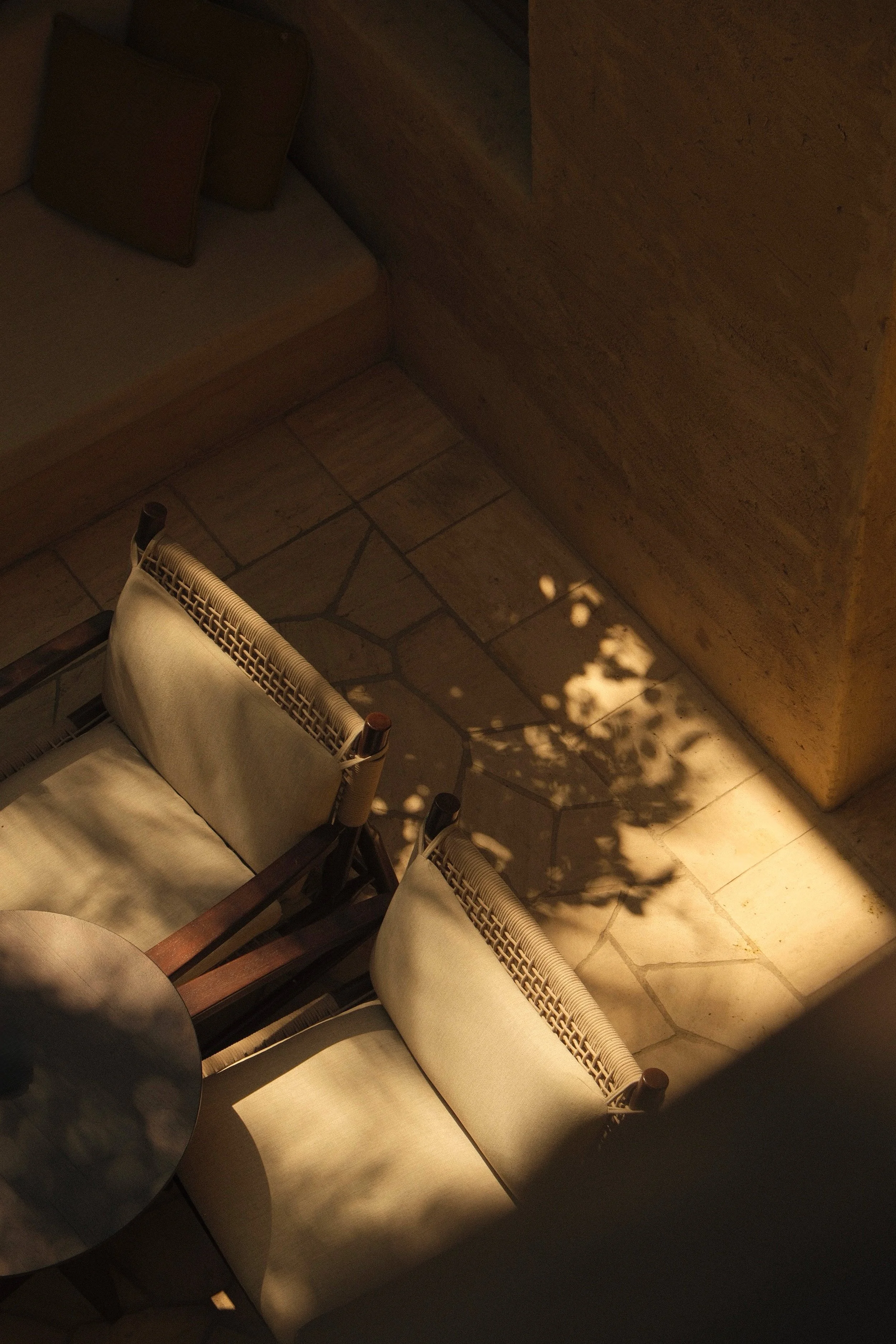 A dimly lit corner of a room showing a wooden chair with beige cushion, a small dark round table, beige tiled floor, and a wooden wall with shadows of tree branches.