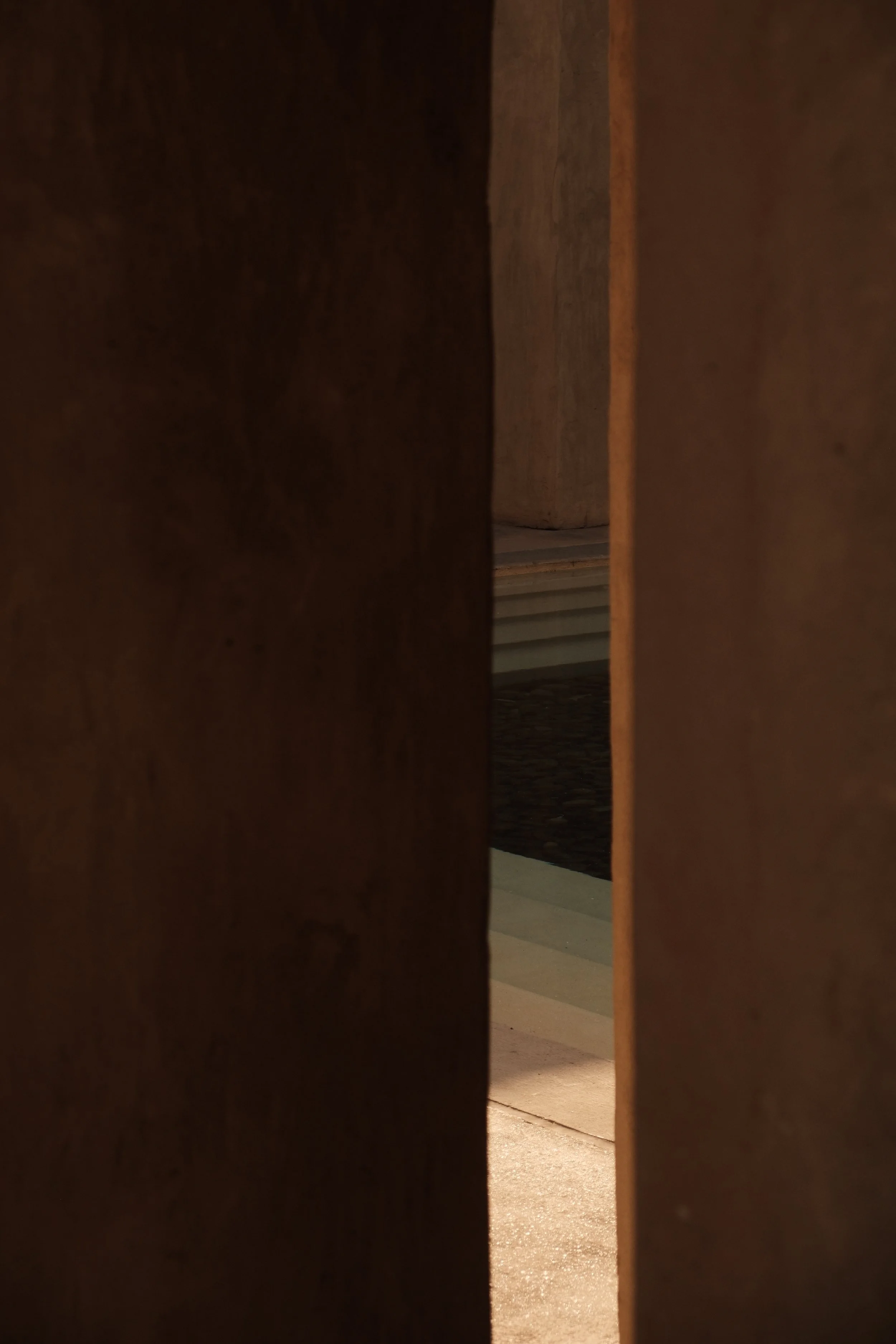 View through a small opening between two wooden panels, showing a space with a floor made of concrete and a dark-colored rug. The background includes a wall and stairs.