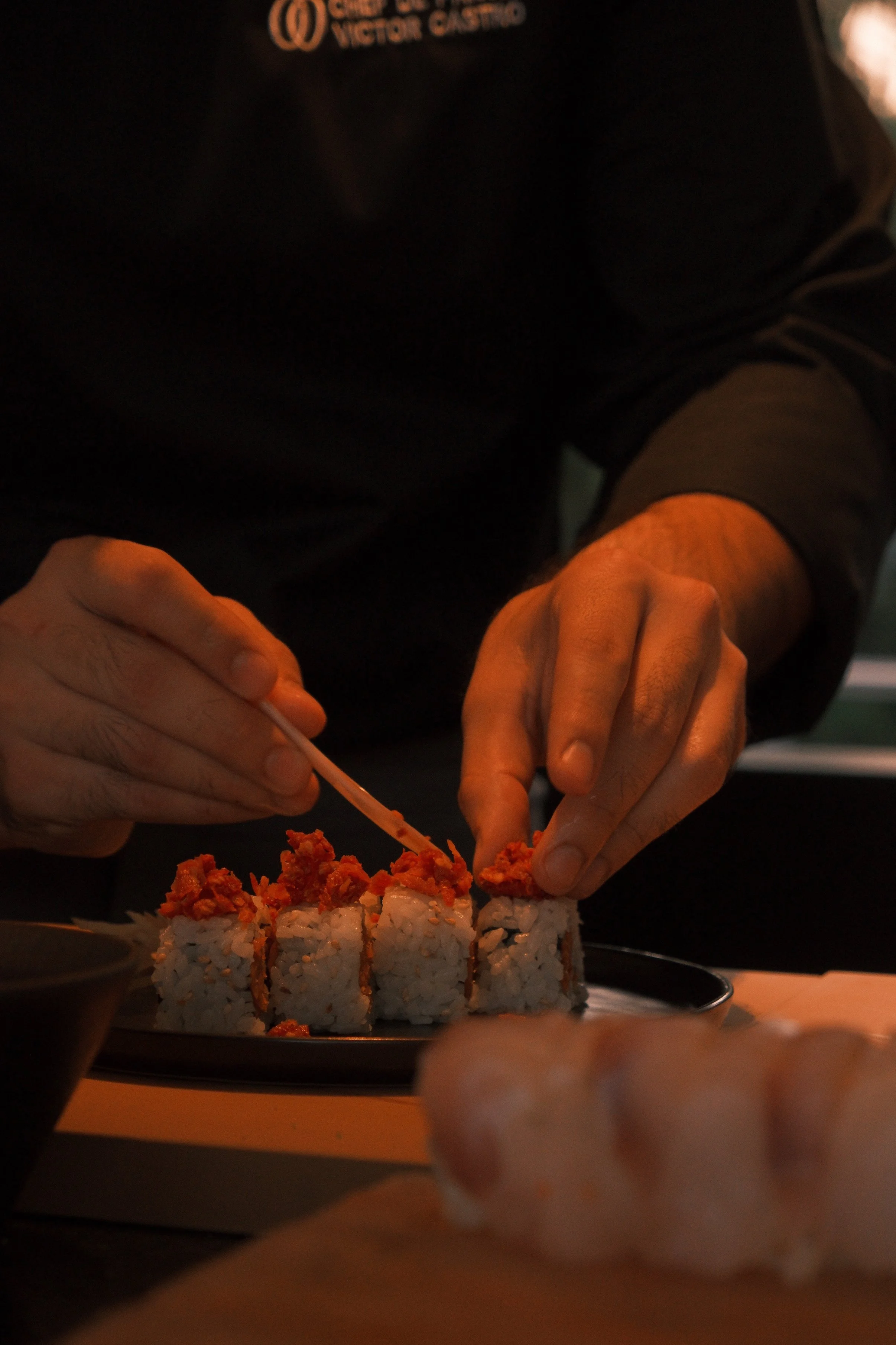 A person preparing sushi rolls, with a focus on their hands as they position the sushi on a black plate. The sushi is topped with spicy mayo and fish roe. The person is wearing a black shirt with an orange logo.