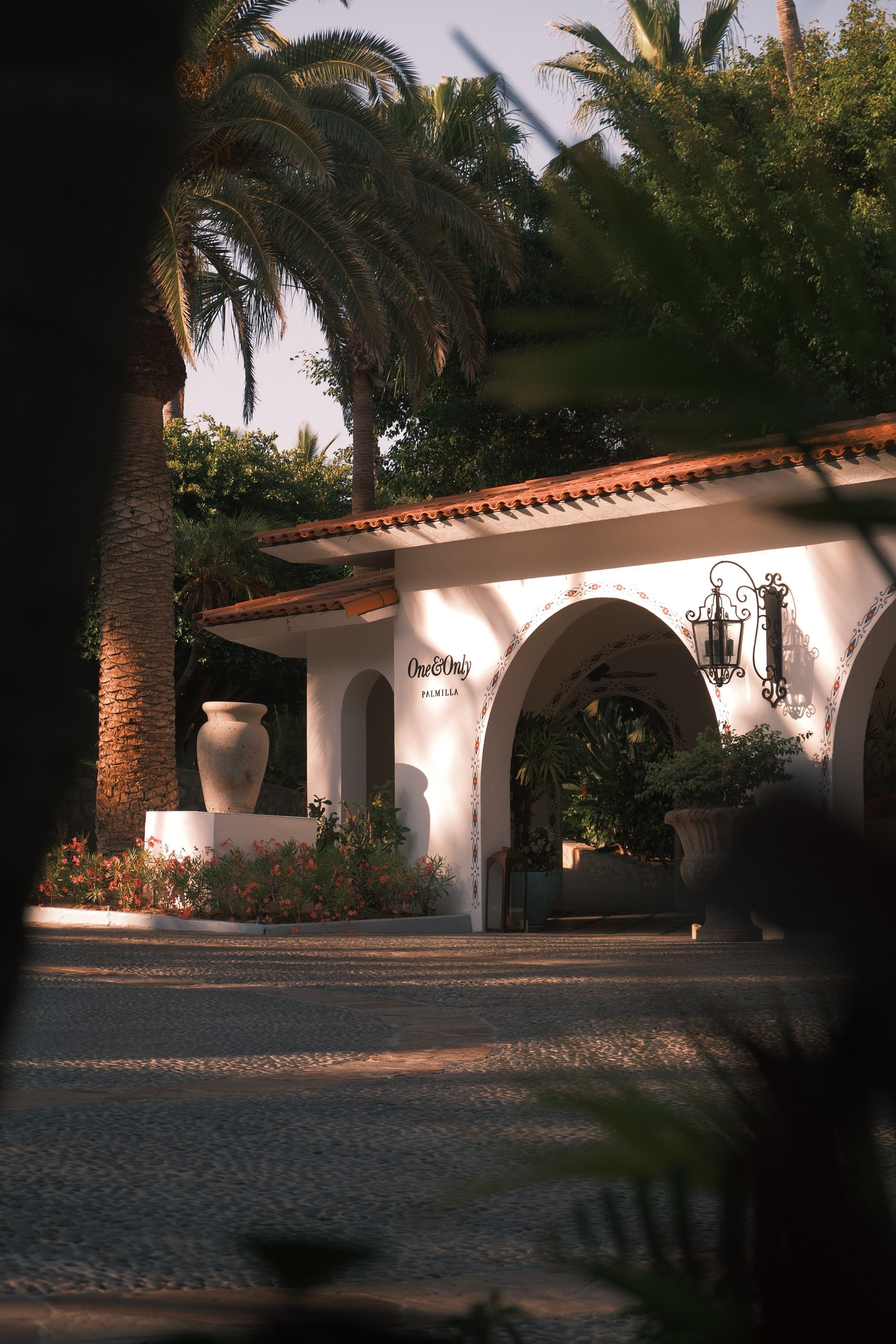 Sunset view of a Mediterranean style building with white walls, red tile roof, decorative lamps, surrounded by palm trees and lush greenery.