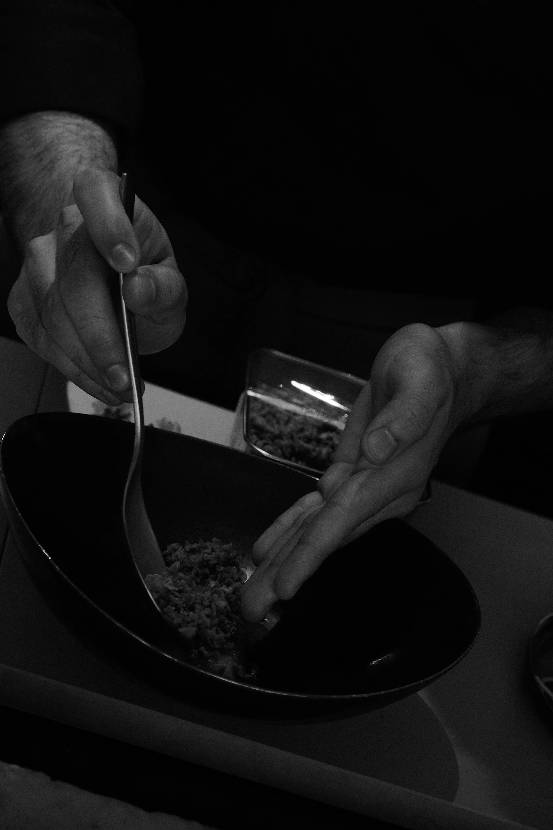 A person is using a fork to serve food from a bowl, with another hand holding a plate. The image is in black and white.
