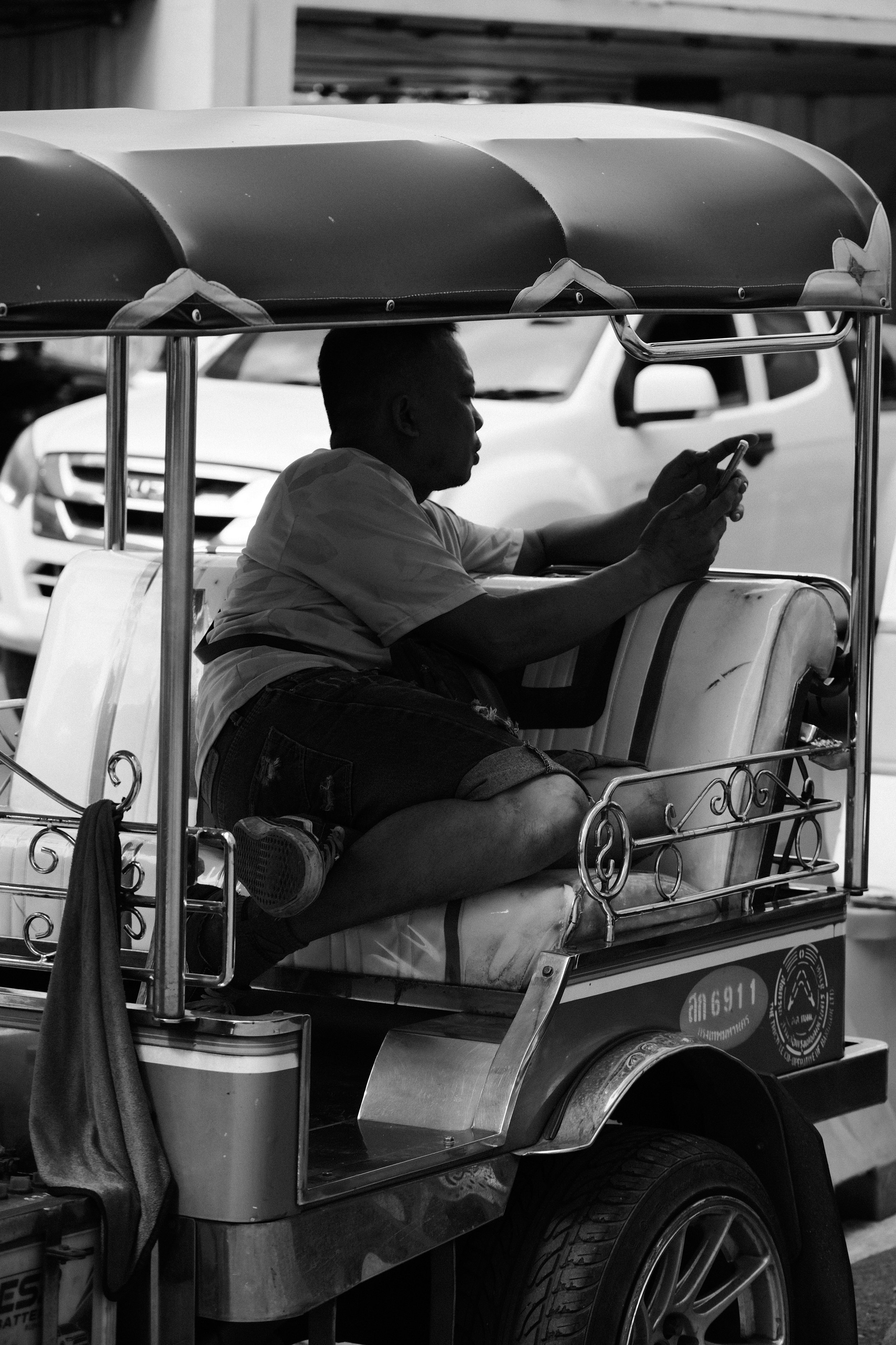 A man sitting in a decorated auto rickshaw, looking at his phone, with a white vehicle and a building in the background.