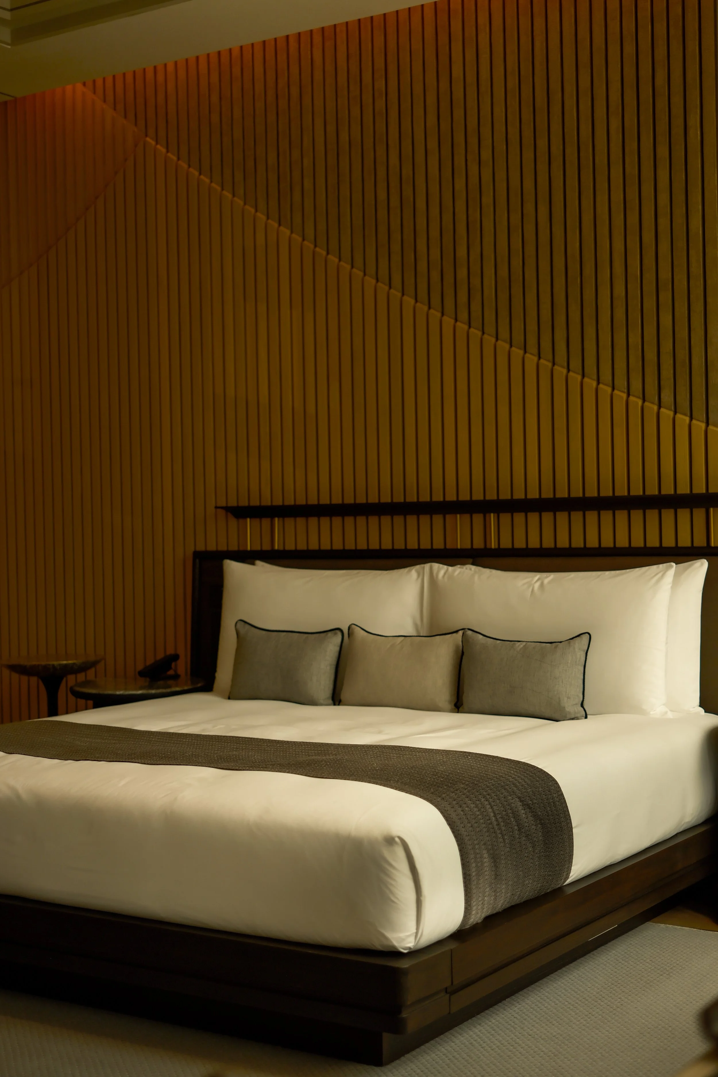 A neatly made bed with white sheets and pillows, a gray runner, and a wooden headboard set against a wall with wood paneling in a modern hotel room.