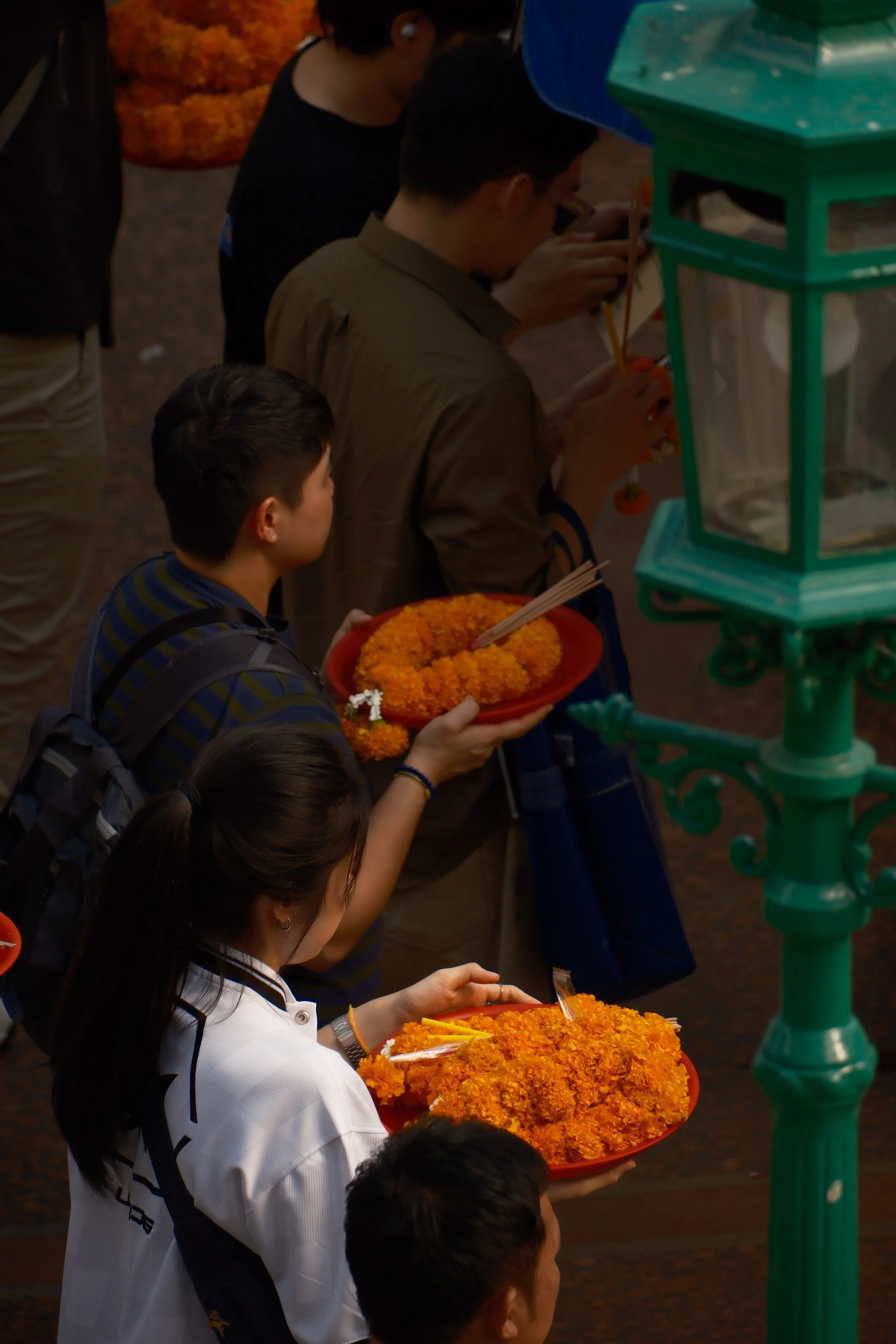 People holding trays of orange marigold flower garlands at a daytime event, standing in line near a green structure.