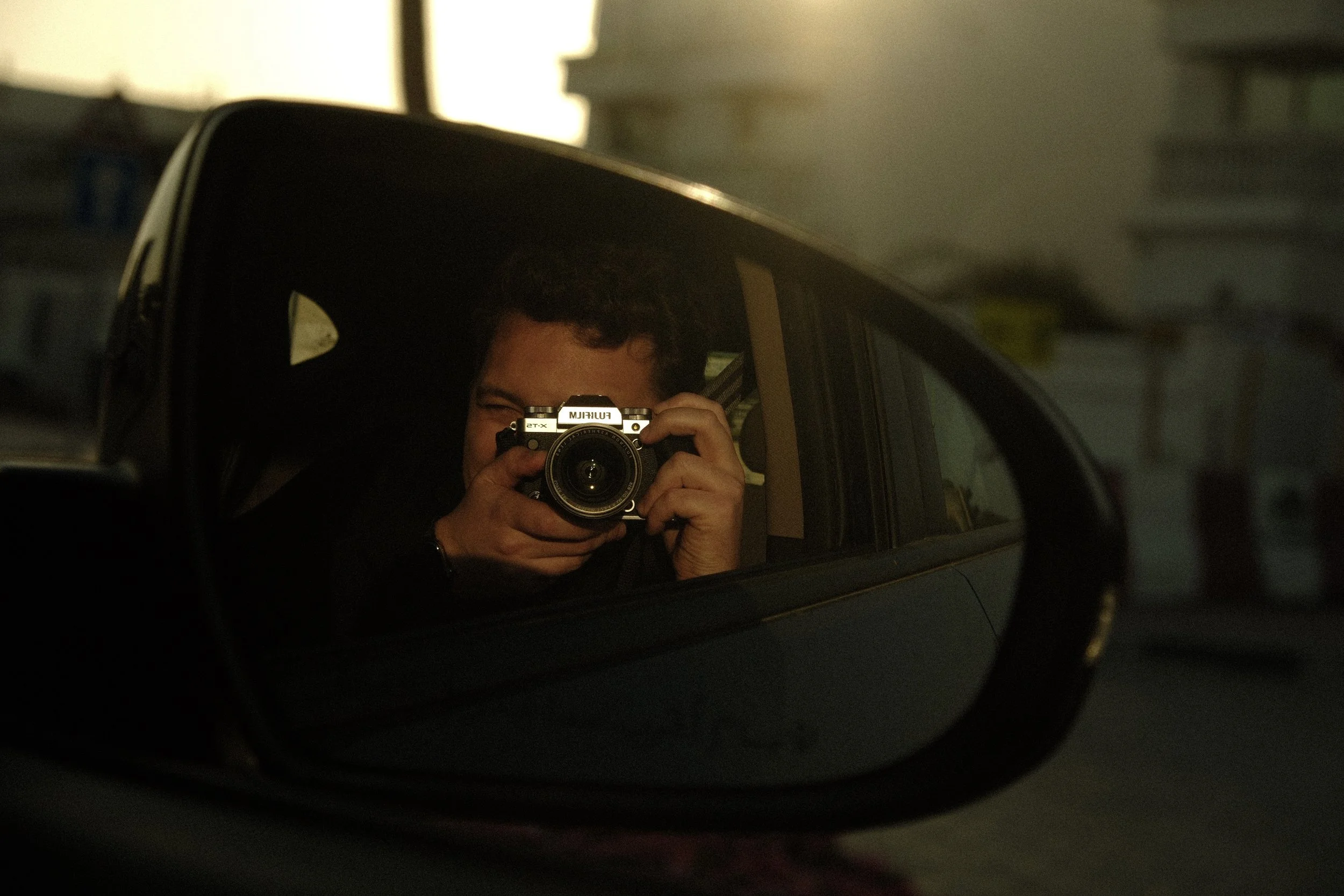 Person taking a photo of themselves in a car's side mirror, with the mirror's reflection capturing their face and camera.