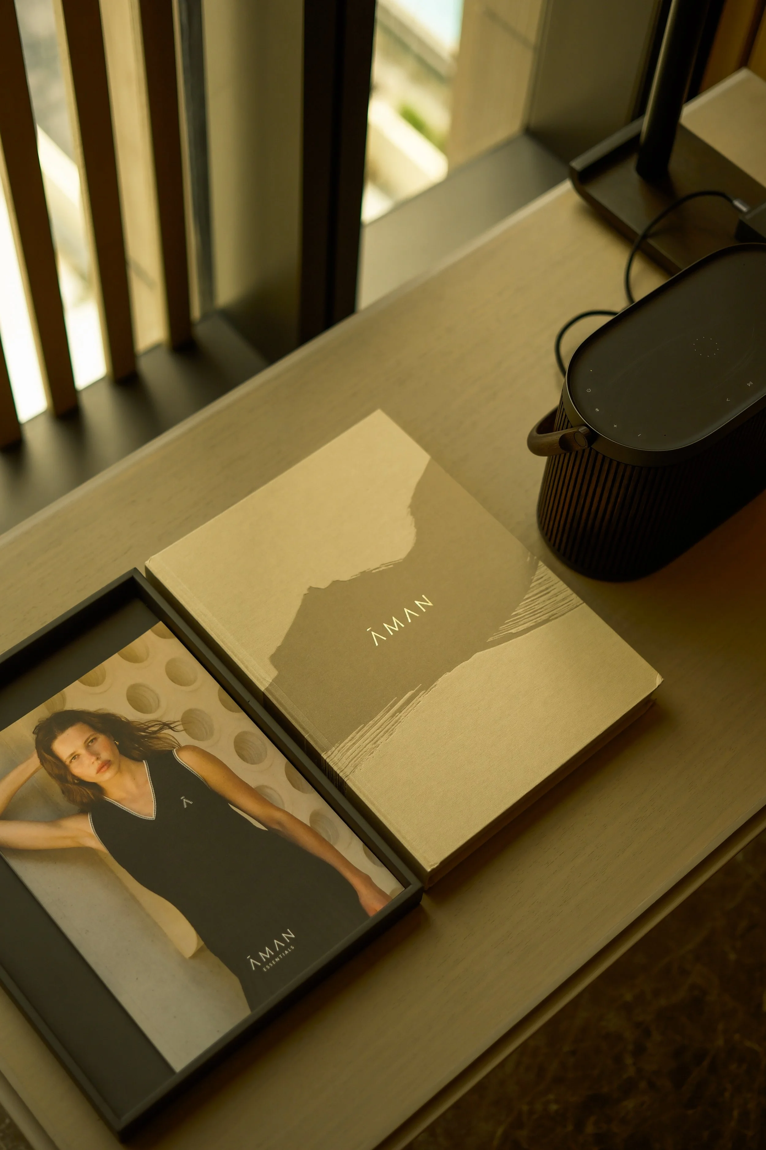 A book titled 'ÁMAN' placed on a table, next to a framed photo of a woman in a black sports top, against a backdrop of a window and wooden blinds.