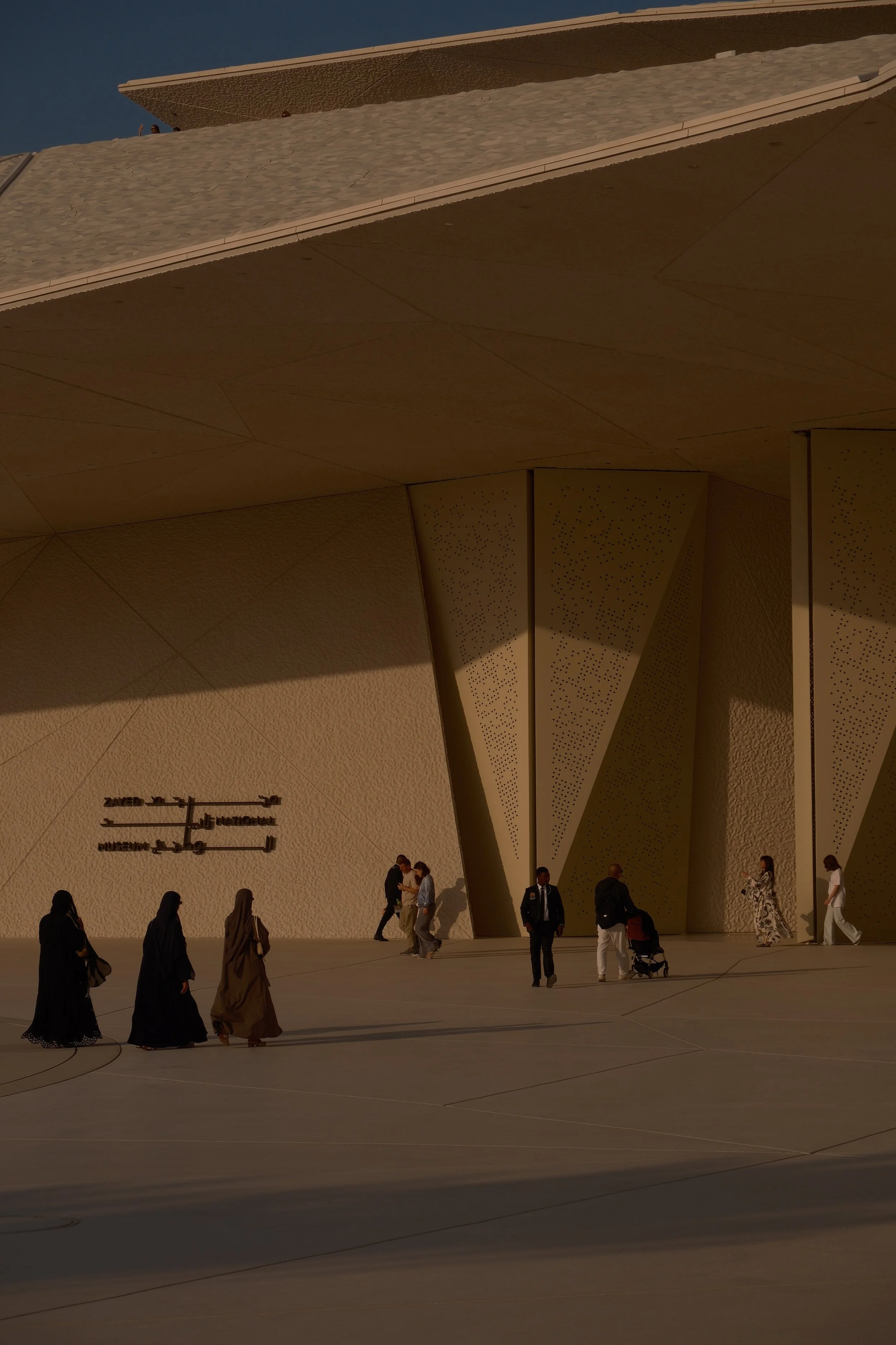 People walking and standing outside a modern architectural building with large, angled overhangs and textured surfaces during daytime.