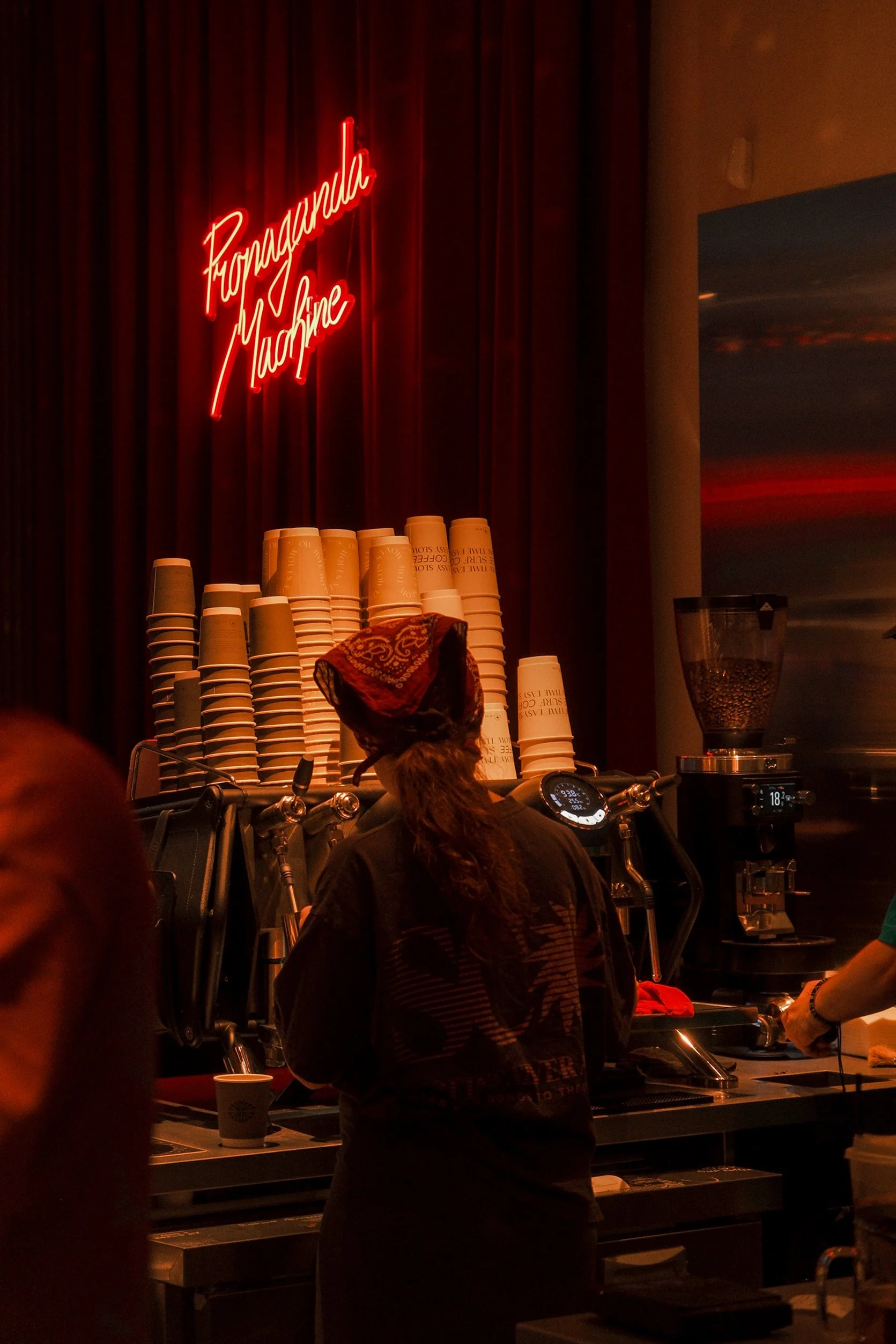 A coffee shop scene with a neon sign reading 'Formaggio Machina' on a dark wood-paneled wall. Several stacks of coffee cups and a barista preparing drinks are visible, with a coffee grinder and part of a menu or equipment in the background.