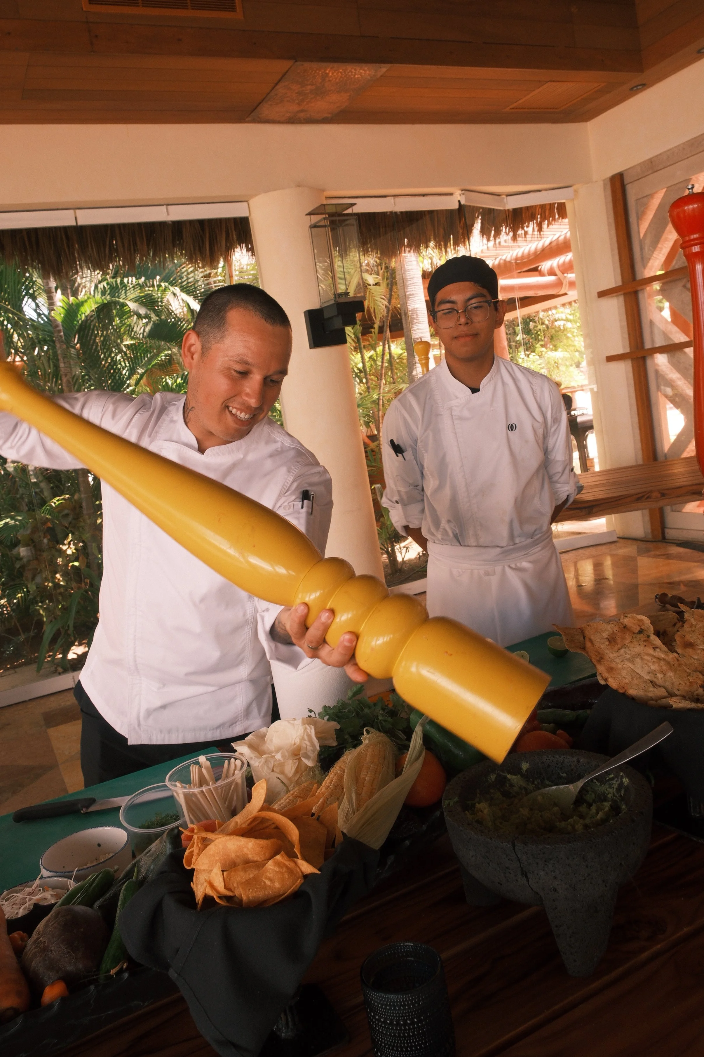 Two chefs in white uniforms preparing food on a table with various ingredients, in a tropical restaurant setting.