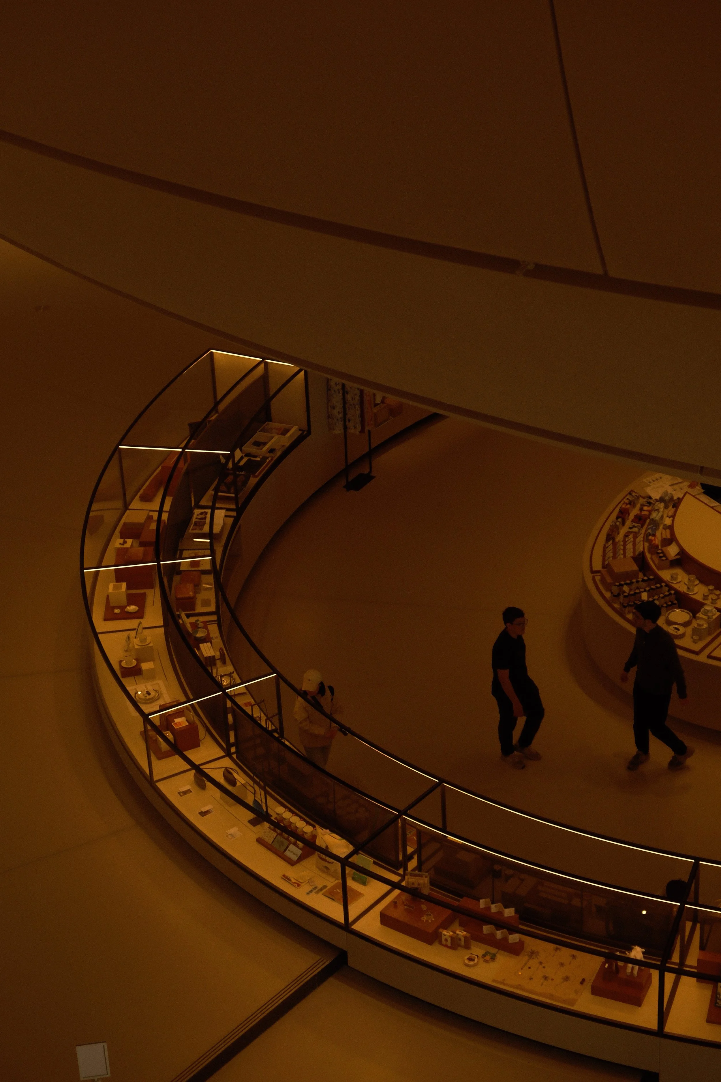 Interior view of a modern museum or gallery with a curved display case filled with various objects and two people walking nearby, with warm lighting.