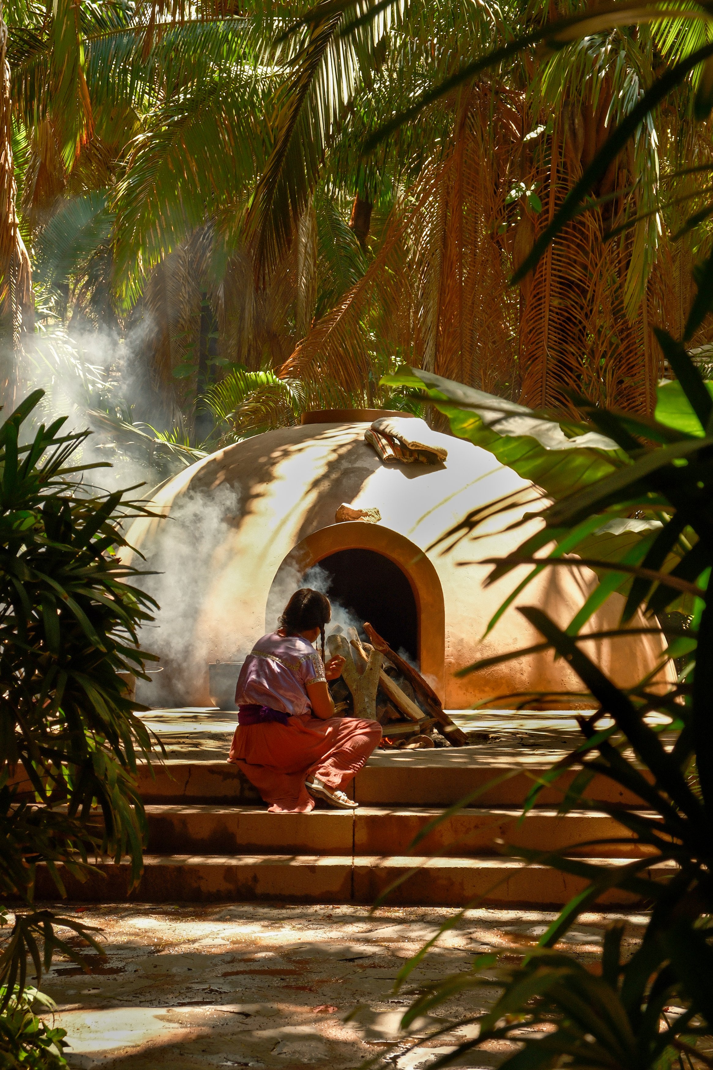 A woman sitting near a clay oven with smoke coming out, surrounded by dense tropical plants and trees.