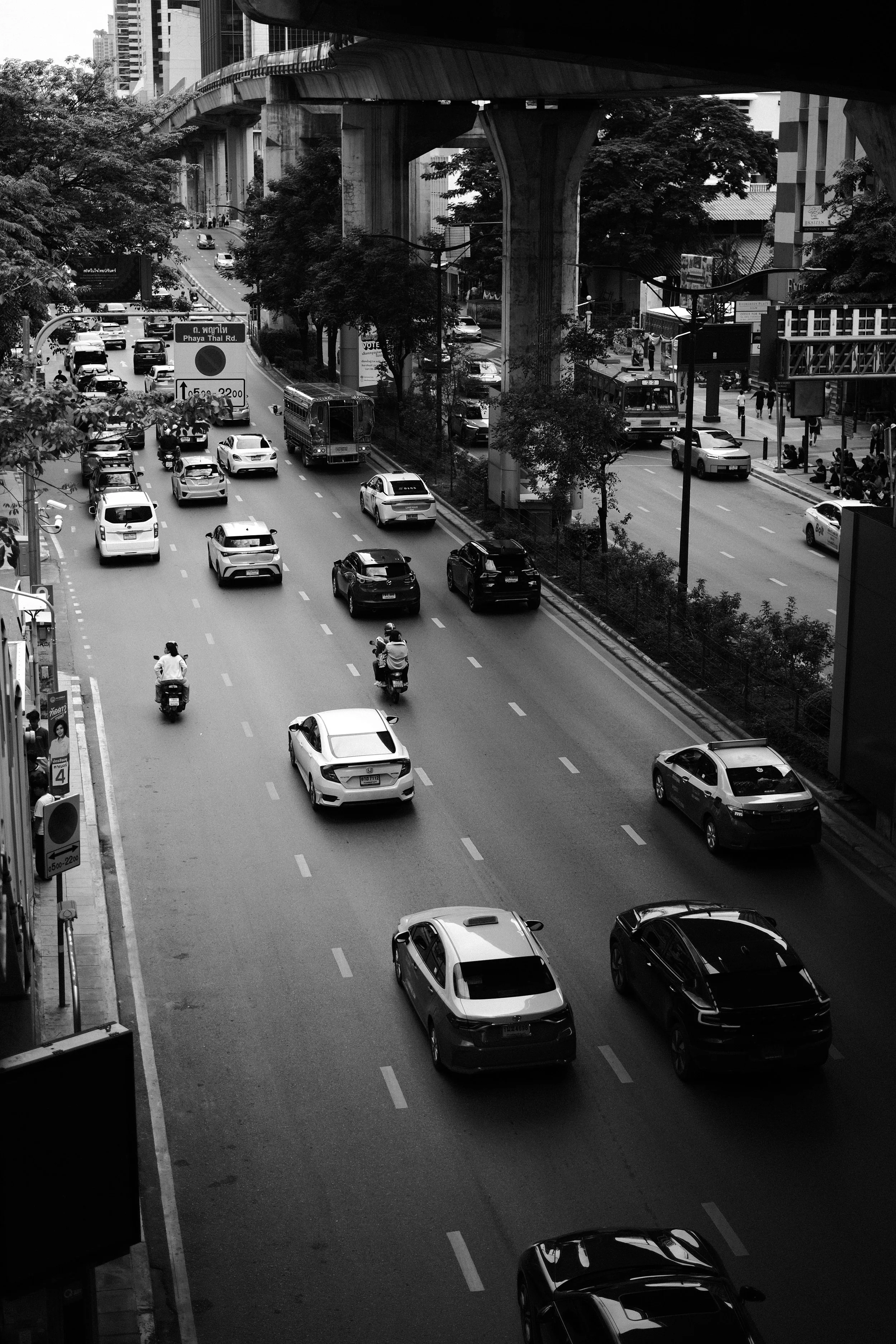 Black-and-white photo of a busy city street with cars and motorcycles, an elevated train track overhead, trees lining the street, and buildings in the background.