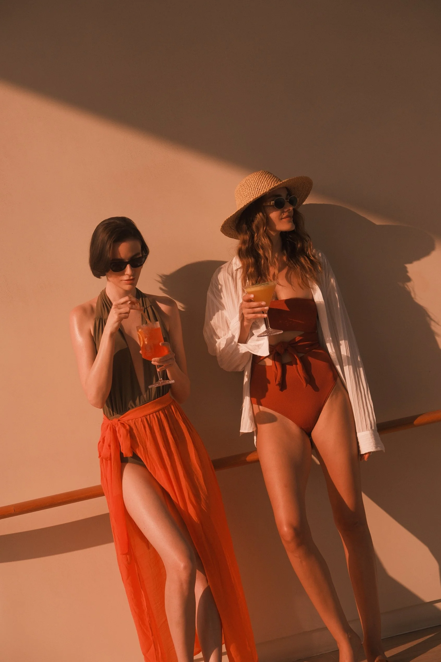 Two women standing against a beige wall, wearing sunglasses, holding drinks, dressed in orange and brown outfits, with sunlight casting shadows on the wall.