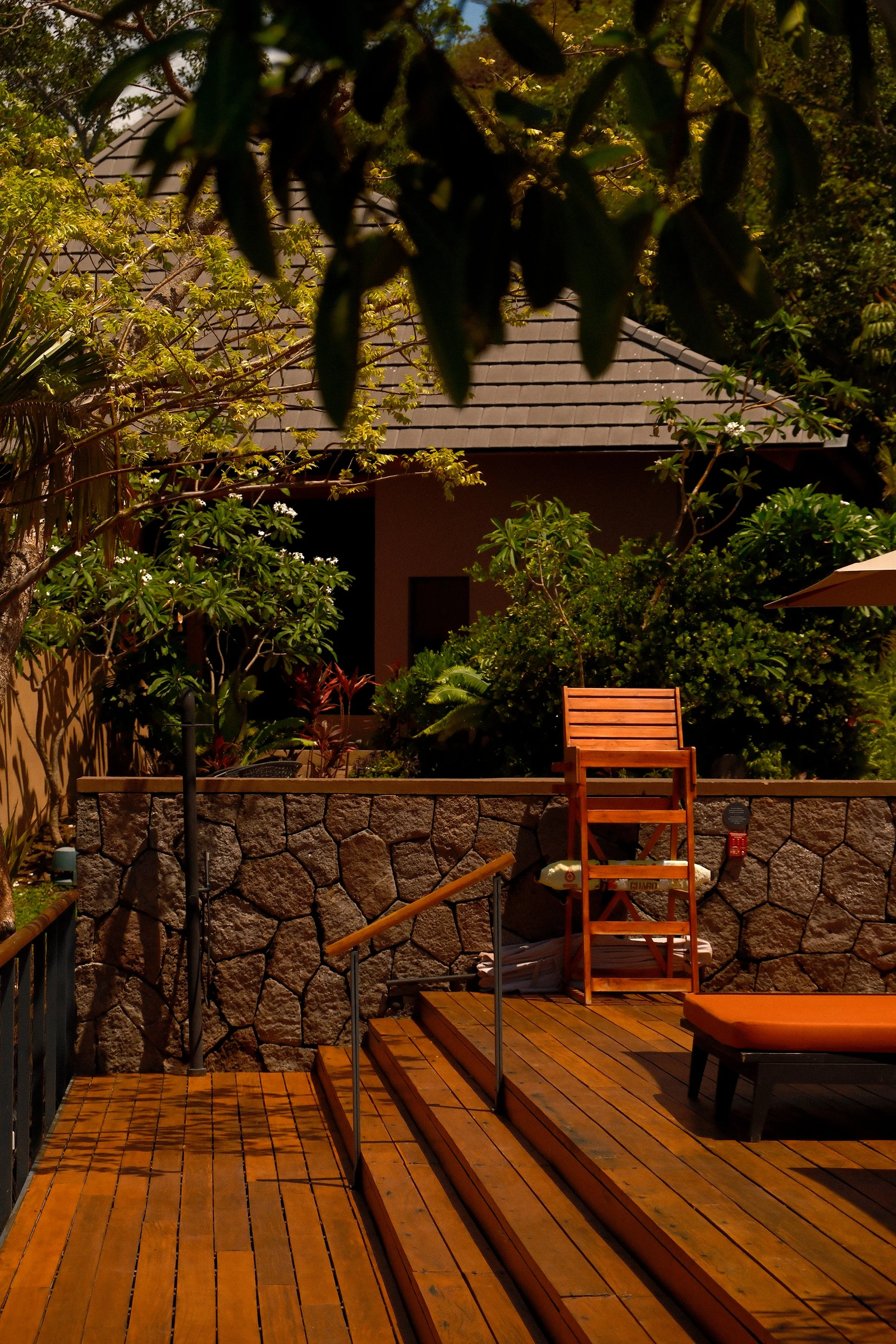 Outdoor patio area at night with wooden deck, steps, a wooden chair, stone wall, lush green plants, trees, and a house with a sloped roof in the background.