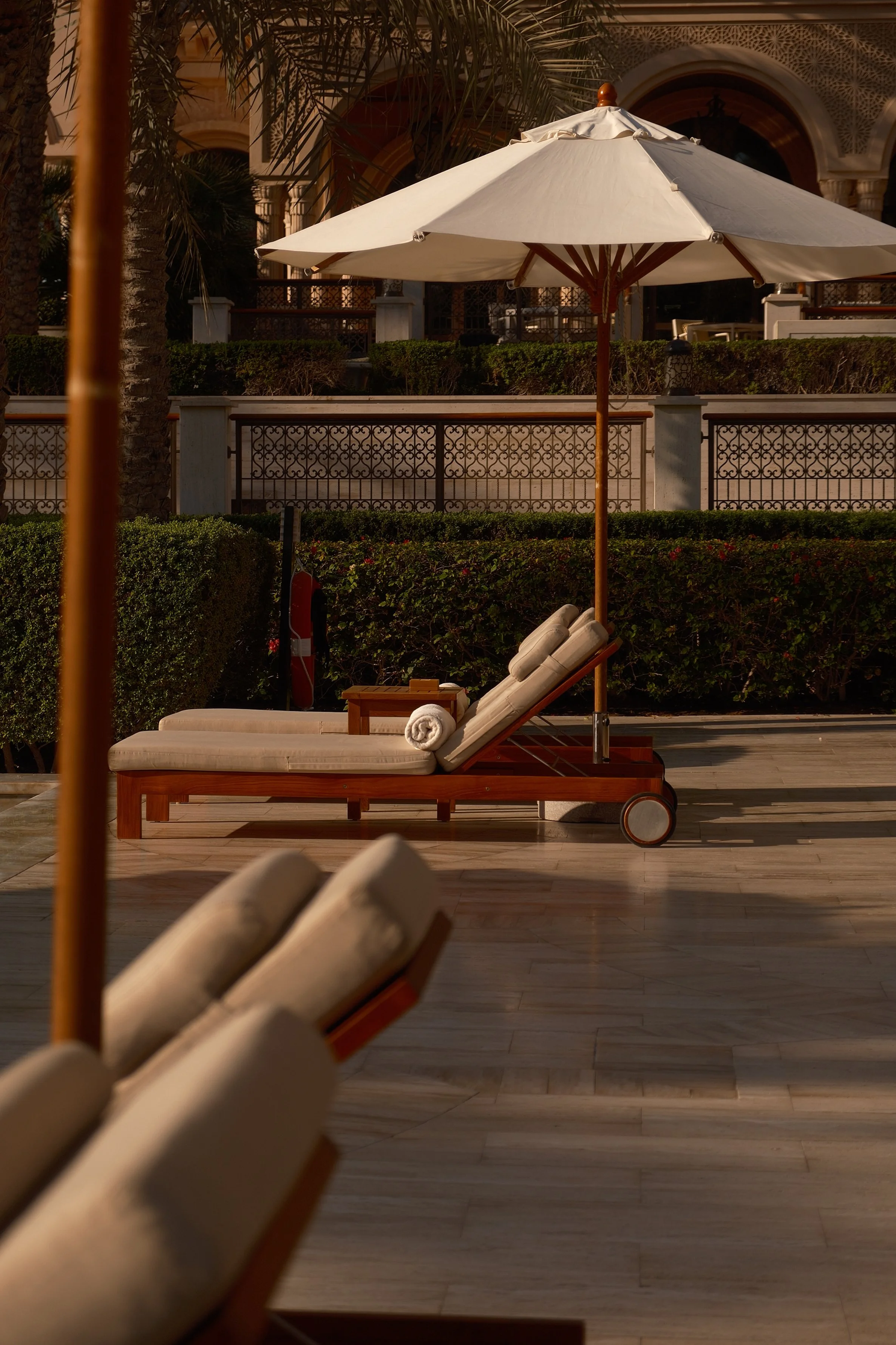 A lounge chair with white cushions and rolled towels sits beneath a large white patio umbrella on a wooden deck. In the background, there is a hedge, a decorative wrought iron fence, palm trees, and a building with ornate arches and detailed decorati