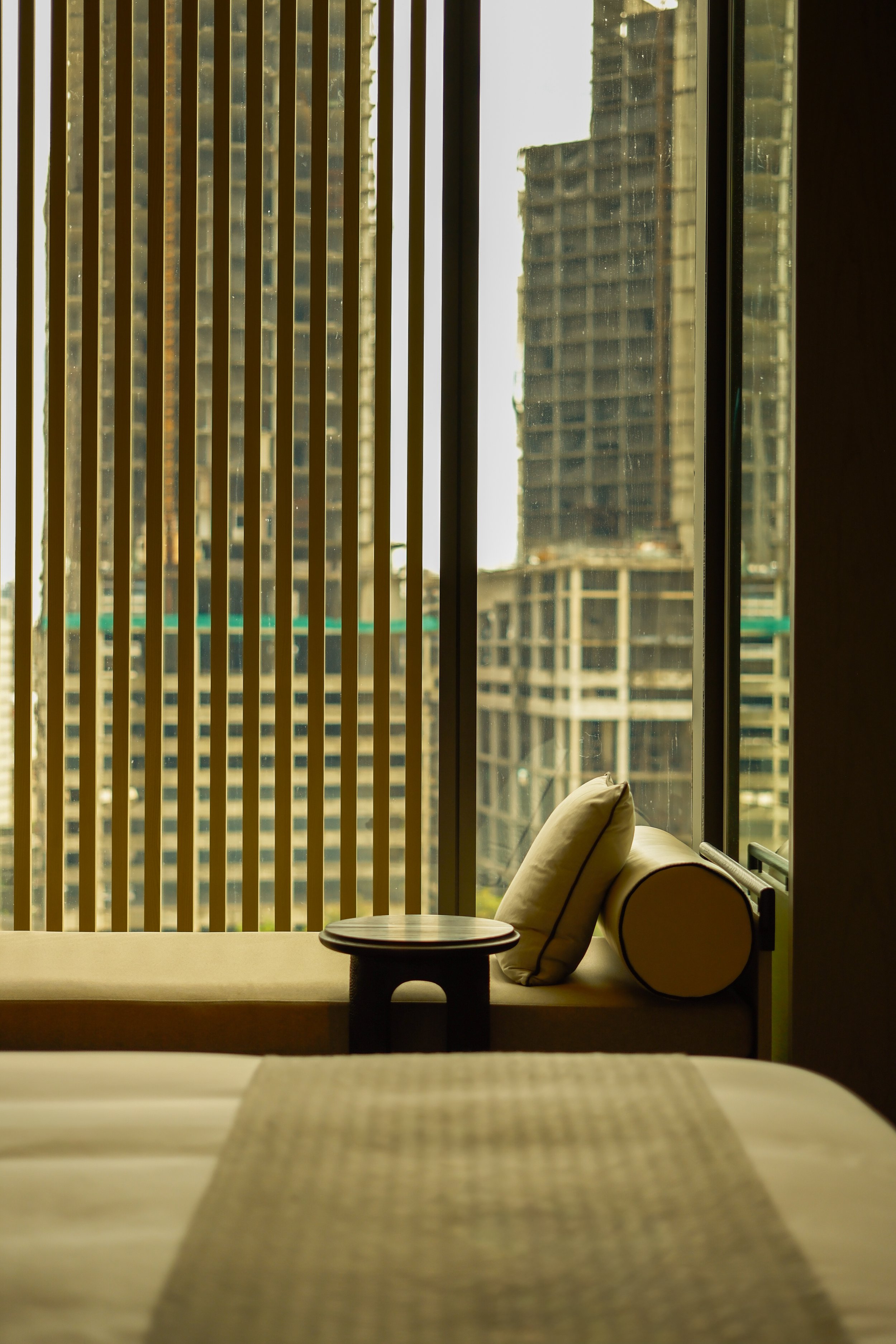Interior of a high-rise apartment with a window seat, plush pillows, a small round table, and vertical blinds, overlooking a cityscape with tall buildings.