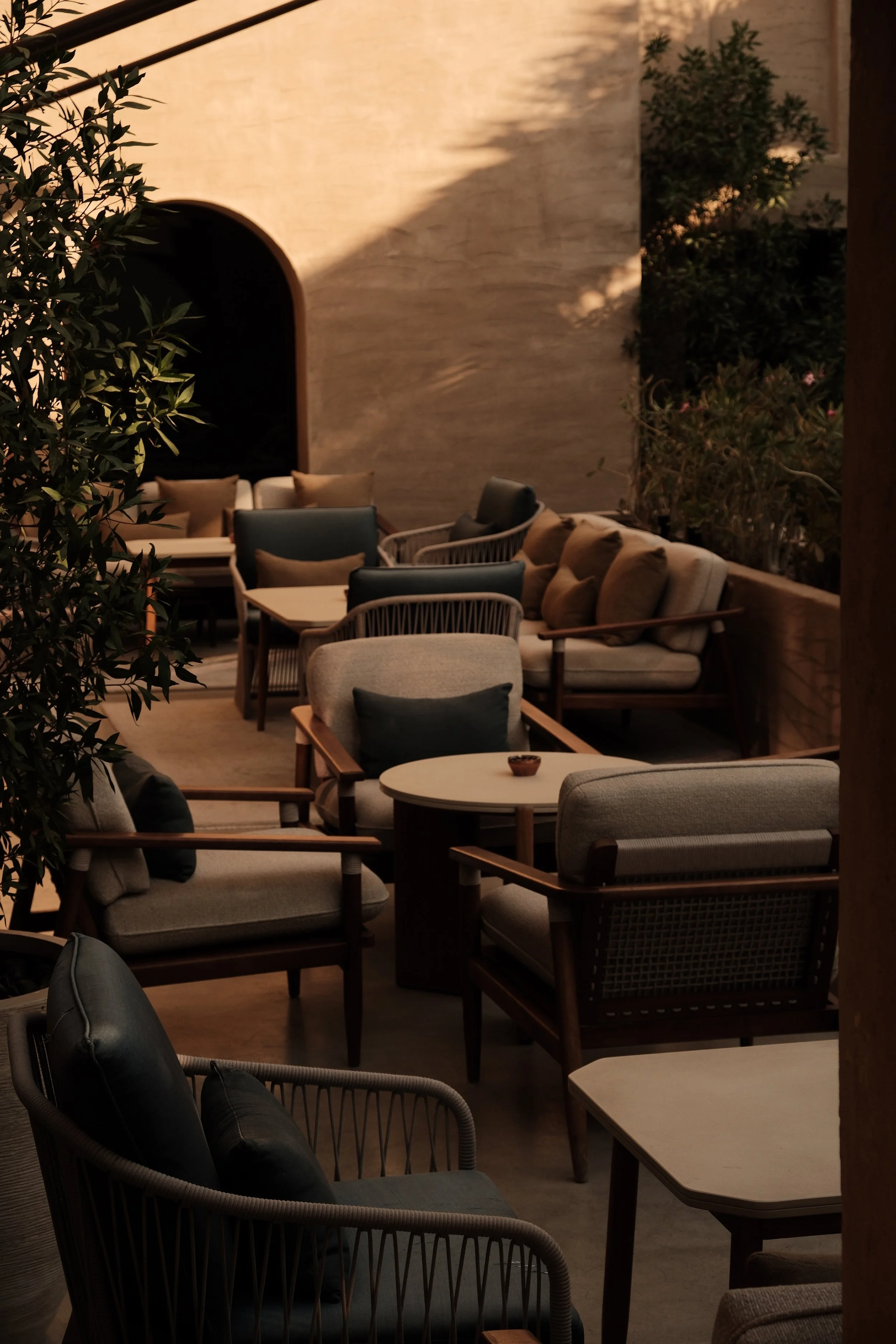 Outdoor seating area with chairs and sofas, some with cushions, in front of a textured wall and surrounded by green plants in a cozy, ambient setting.
