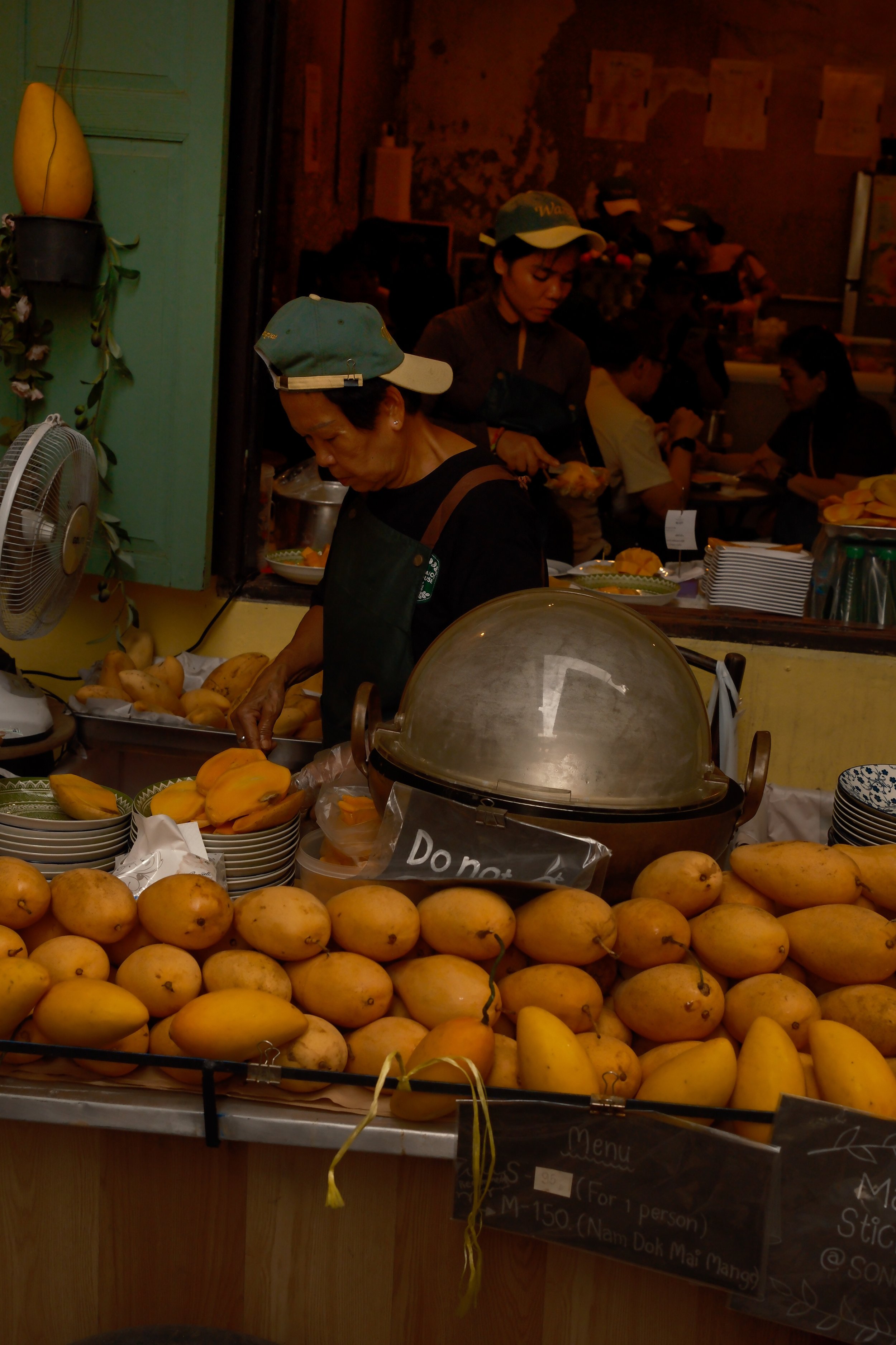 Fresh yellow mangoes arranged on a stall counter at a market, with a woman preparing them and various bowls and signs in the background.