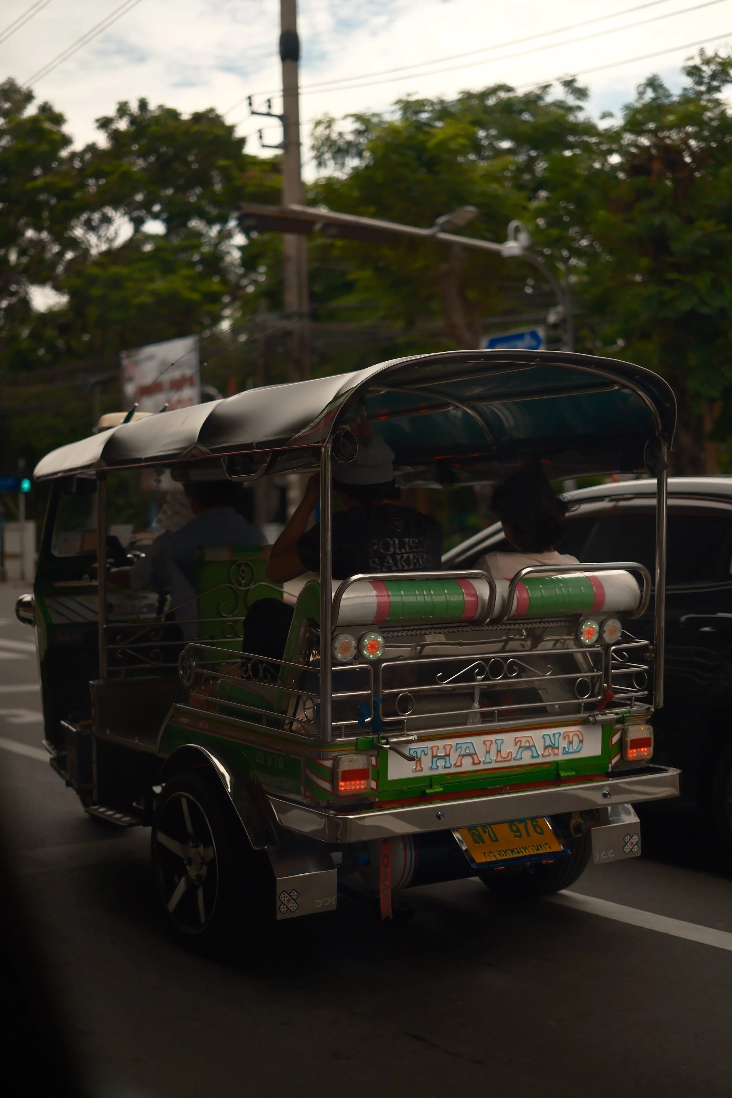 Colorful tuk-tuk with 'Thailand' written on the back, driving on a street with trees and utility poles in the background.