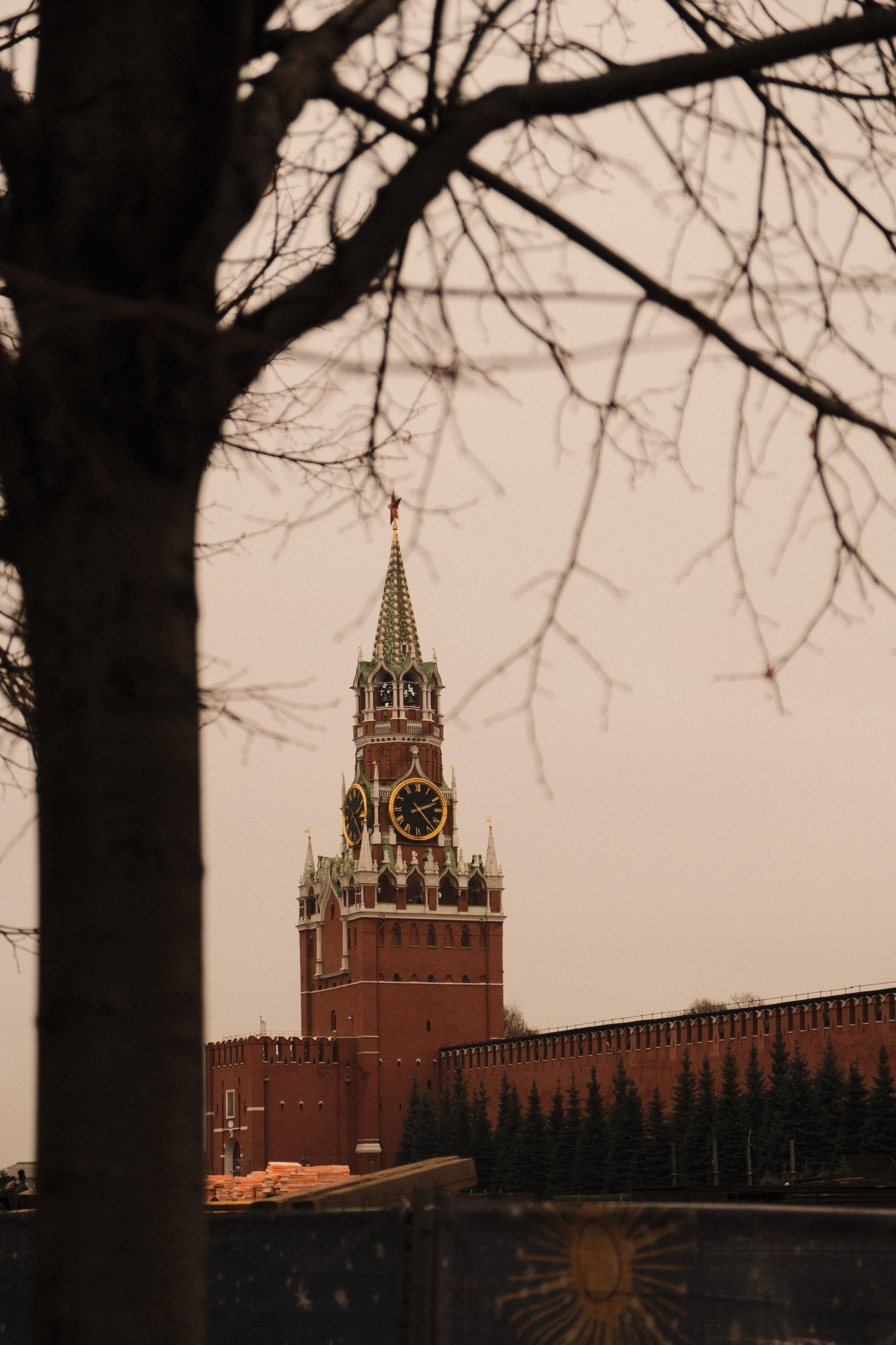 The iconic Spasskaya Tower of the Moscow Kremlin with a clock, viewed through leafless tree branches against an overcast sky.