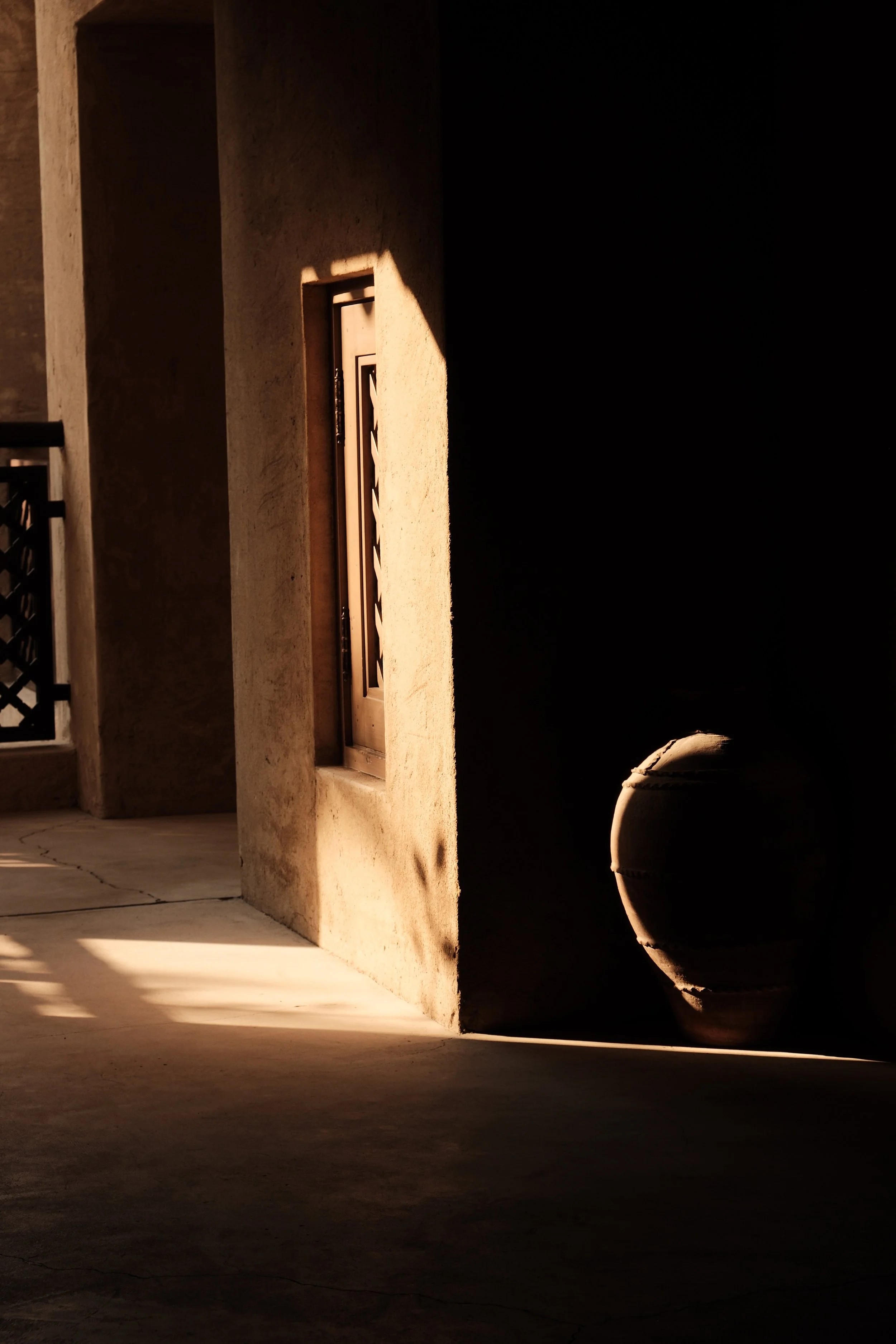 A shadowed outdoor scene with a beige wall, a small vent, and a large clay pot, with sunlight casting shadows on the ground and wall.