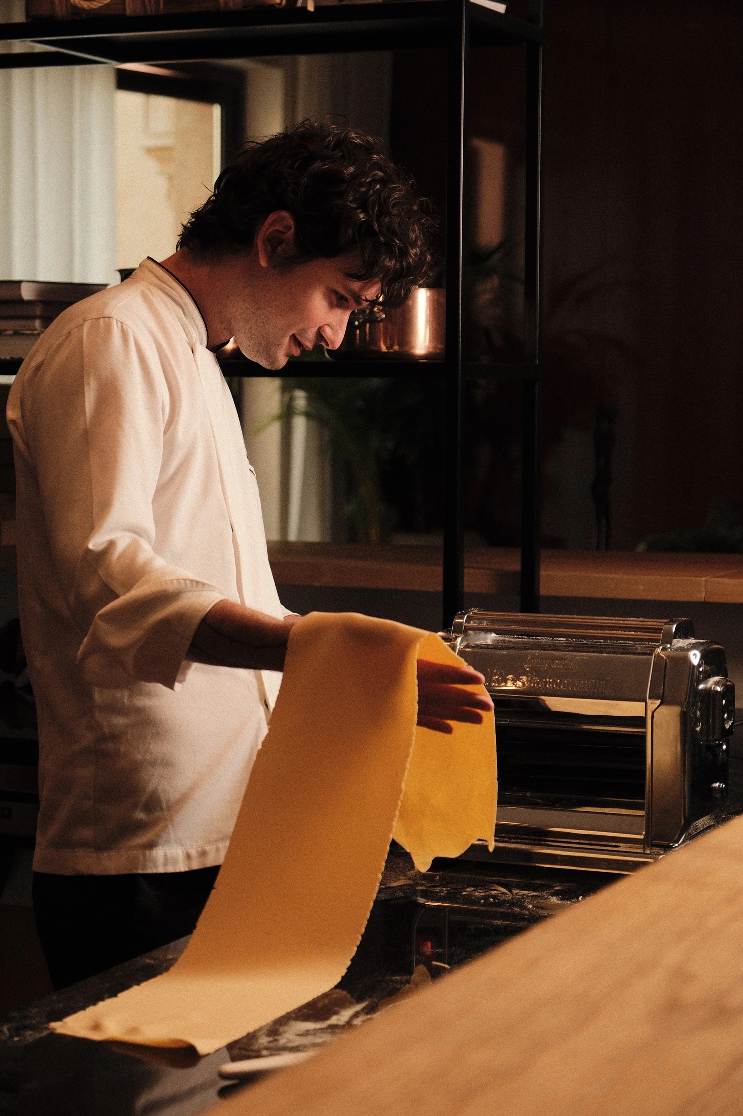 A chef wearing a white uniform and yellow oven mitts is preparing fresh pasta dough in a restaurant kitchen. There is a pasta machine on the counter and industrial shelving in the background.