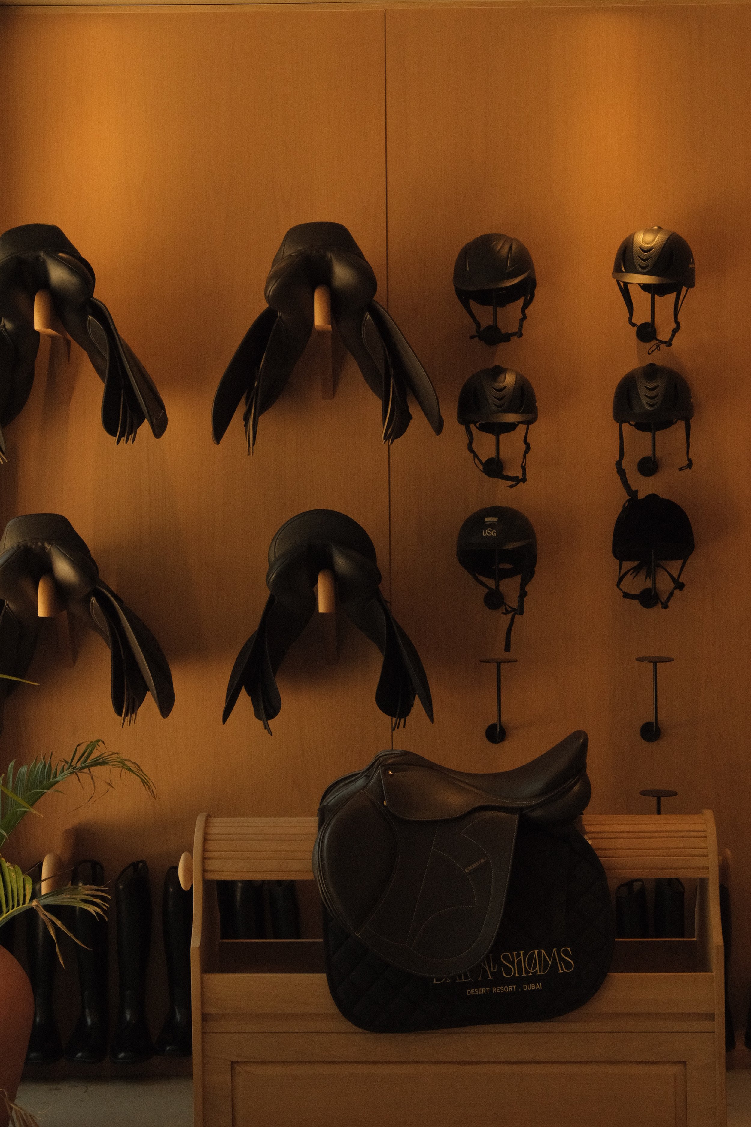 Display of black riding helmets and black riding saddles in a room with wooden furniture and a wooden wall. Saddles are hung on hooks and helmets are on shelves.