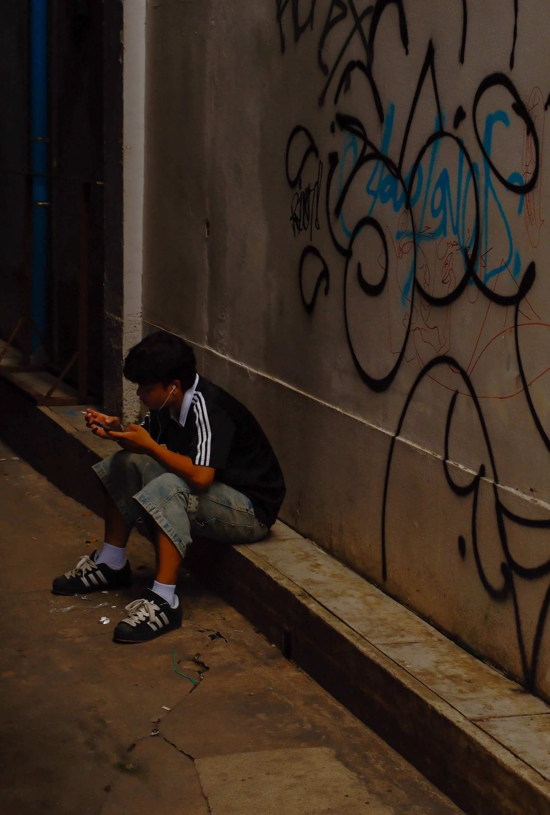 A boy sitting on a sidewalk at night, engrossed in his phone, with black and white sneakers, wearing a black jacket with white stripes on the sleeves and denim shorts. He has dark hair and appears to be using earphones. Behind him is a concrete wall 