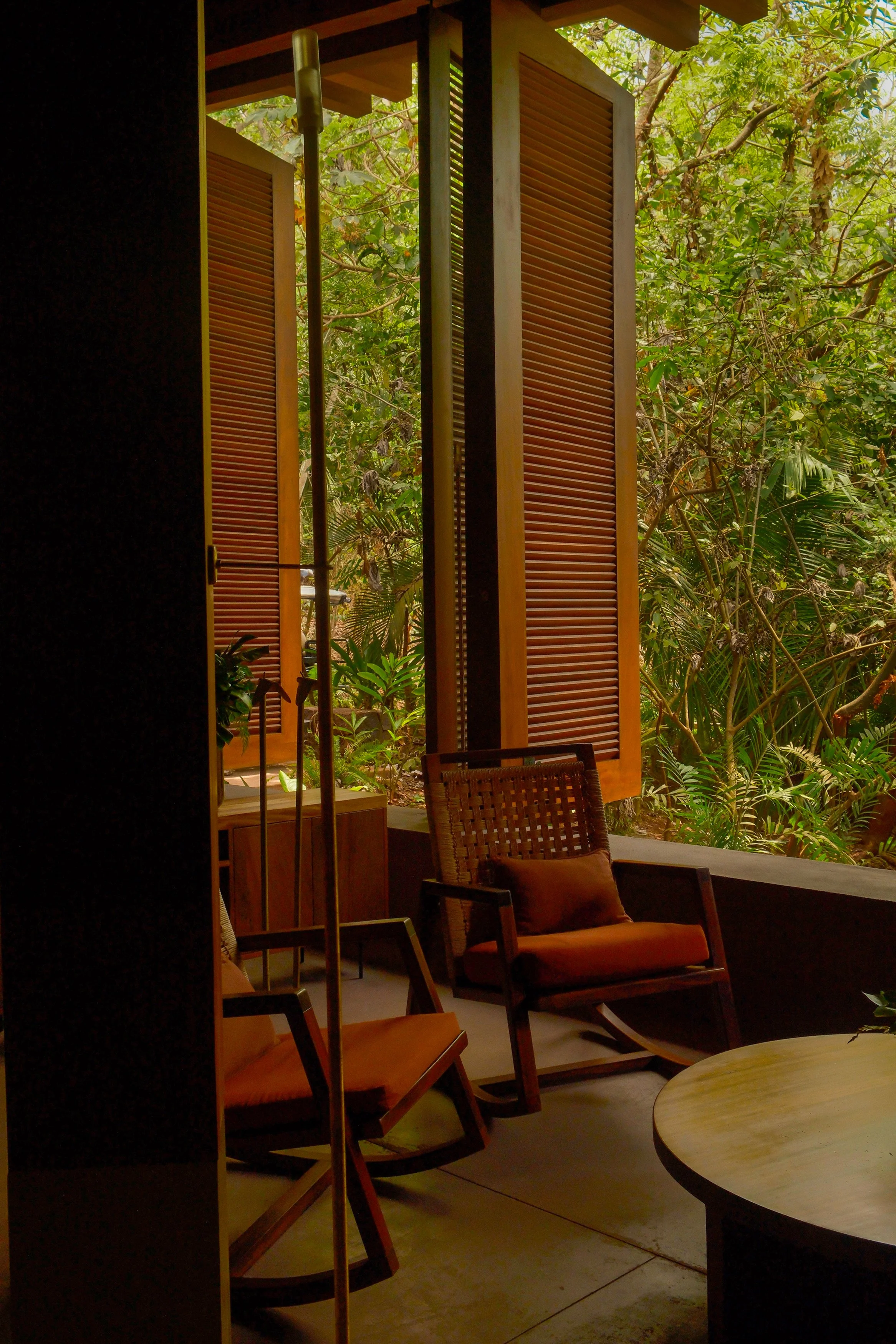 Interior view of a cozy seating area with wooden chairs and a round table, large windows with wooden shutters opening to a lush green jungle outside.