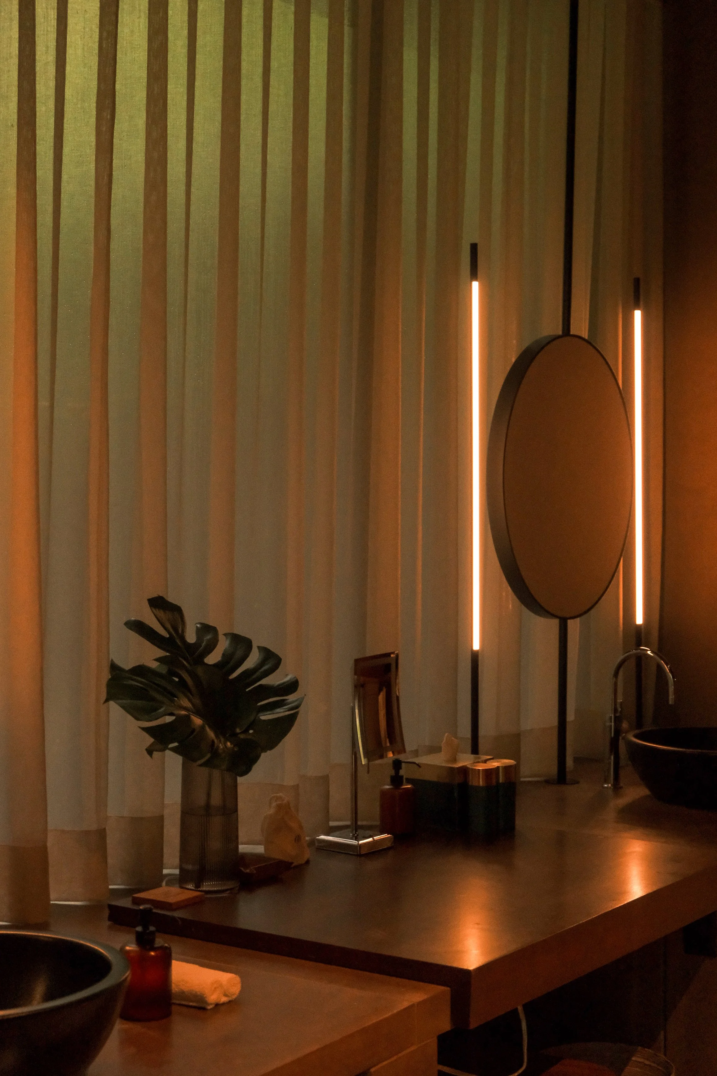 Interior of a modern room with vertical striped curtains, a large round mirror illuminated with vertical light strips, a tall plant in a glass dark vase, and a wooden table with decorative items and a sink nearby.