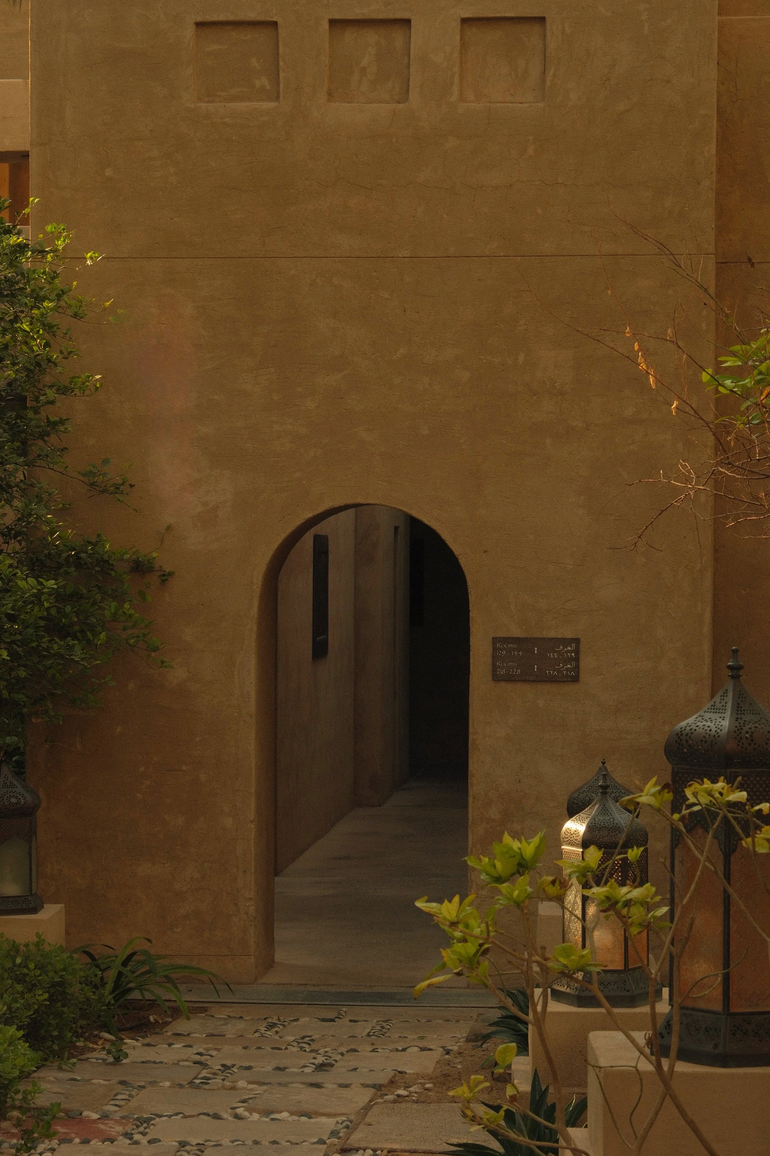 A beige outdoor wall with three square niches near the top, an arched doorway, and a sign with room numbers in English and Arabic on the right. There are green plants and decorative lanterns in the foreground.