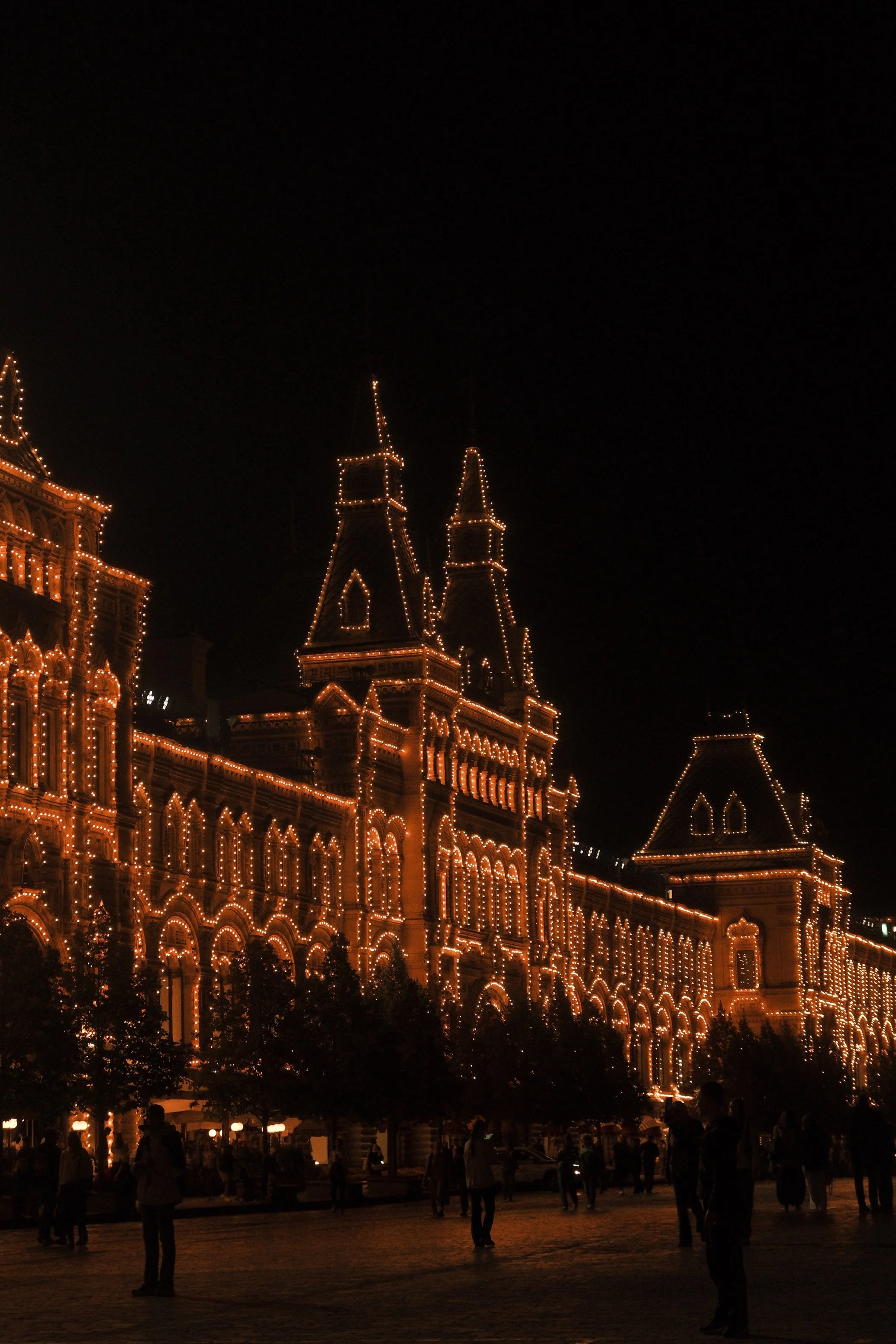 Nighttime view of a large, historic building decorated with Christmas lights, with people walking in the foreground.