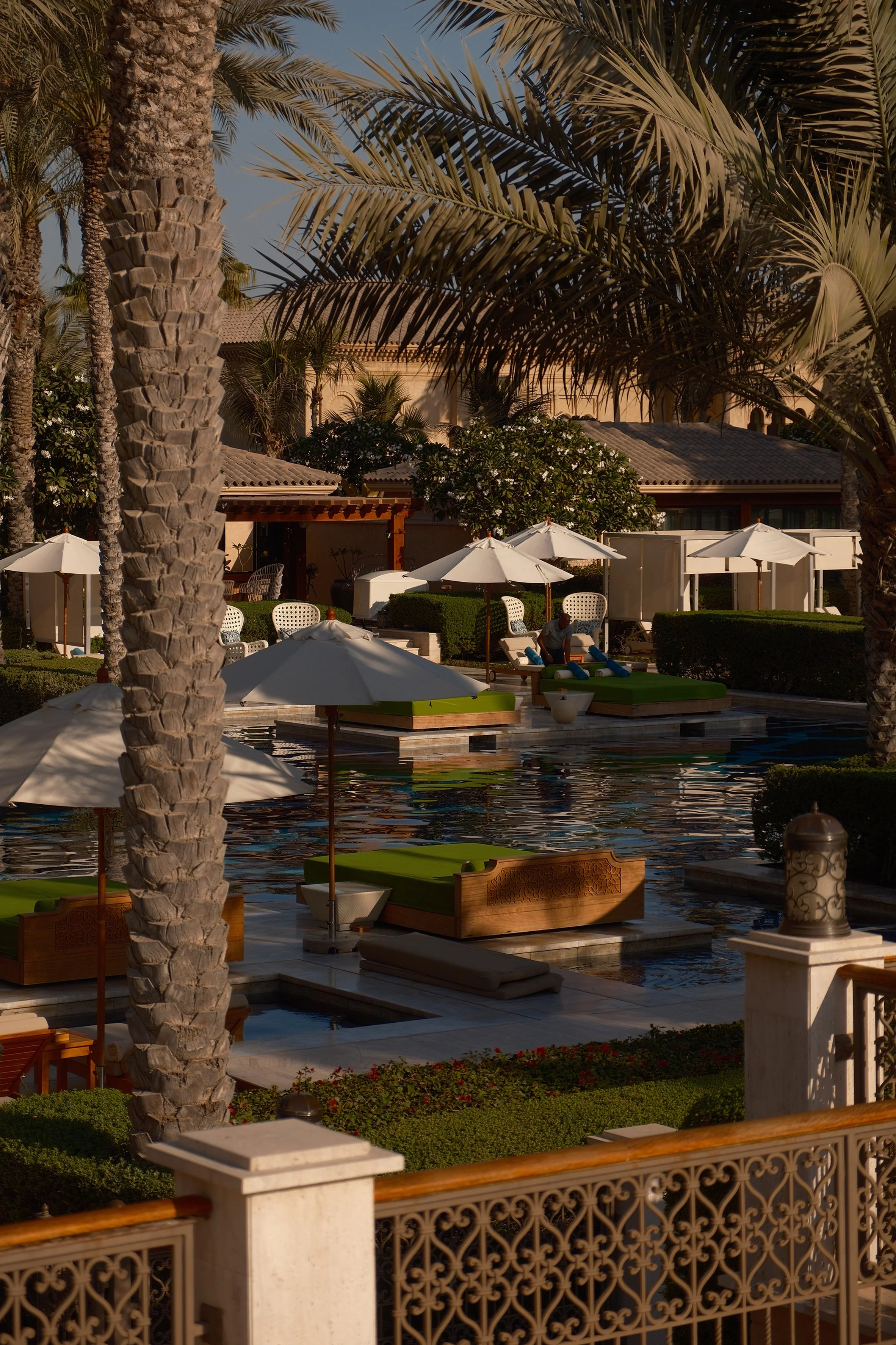 Luxury outdoor pool area with palm trees, white umbrellas, sun loungers, and lush greenery in a sunny setting.