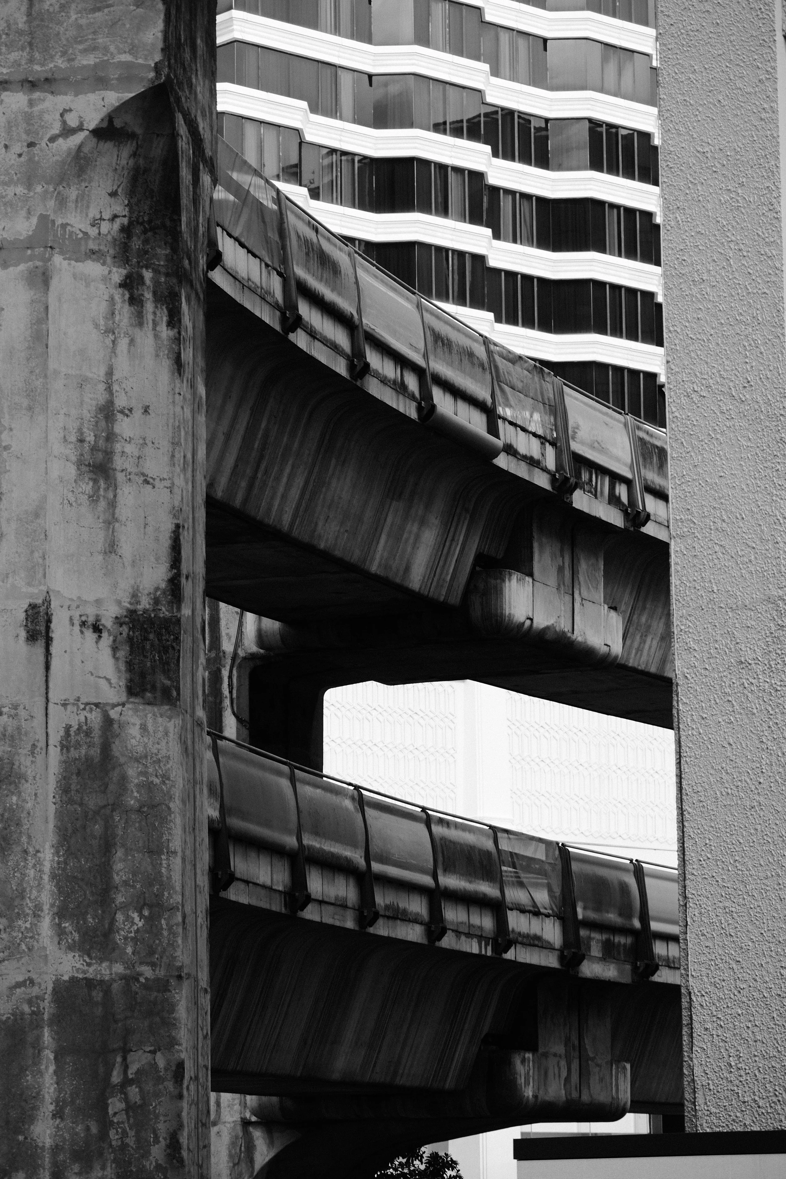 Black and white photograph of a concrete and steel overpass or bridge with a modern multi-story building in the background.
