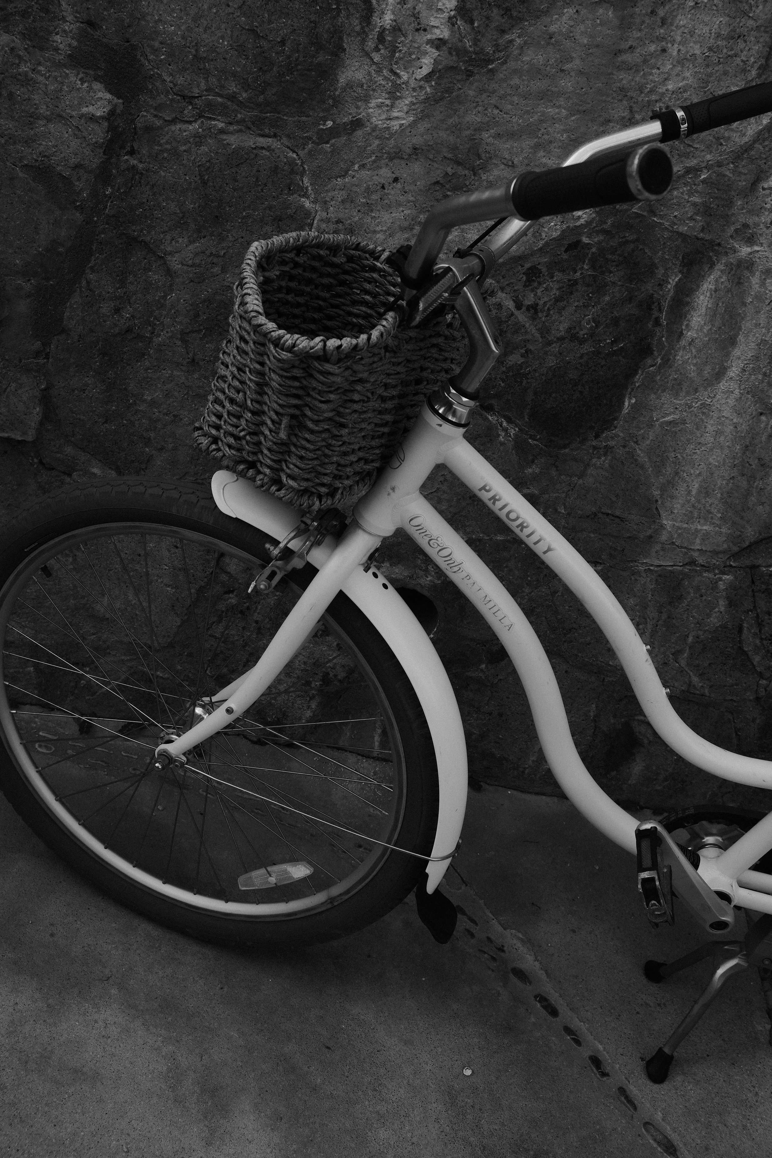 A white bicycle with a wicker basket attached to the front handlebars, leaning against a textured stone wall. The image is in black and white.